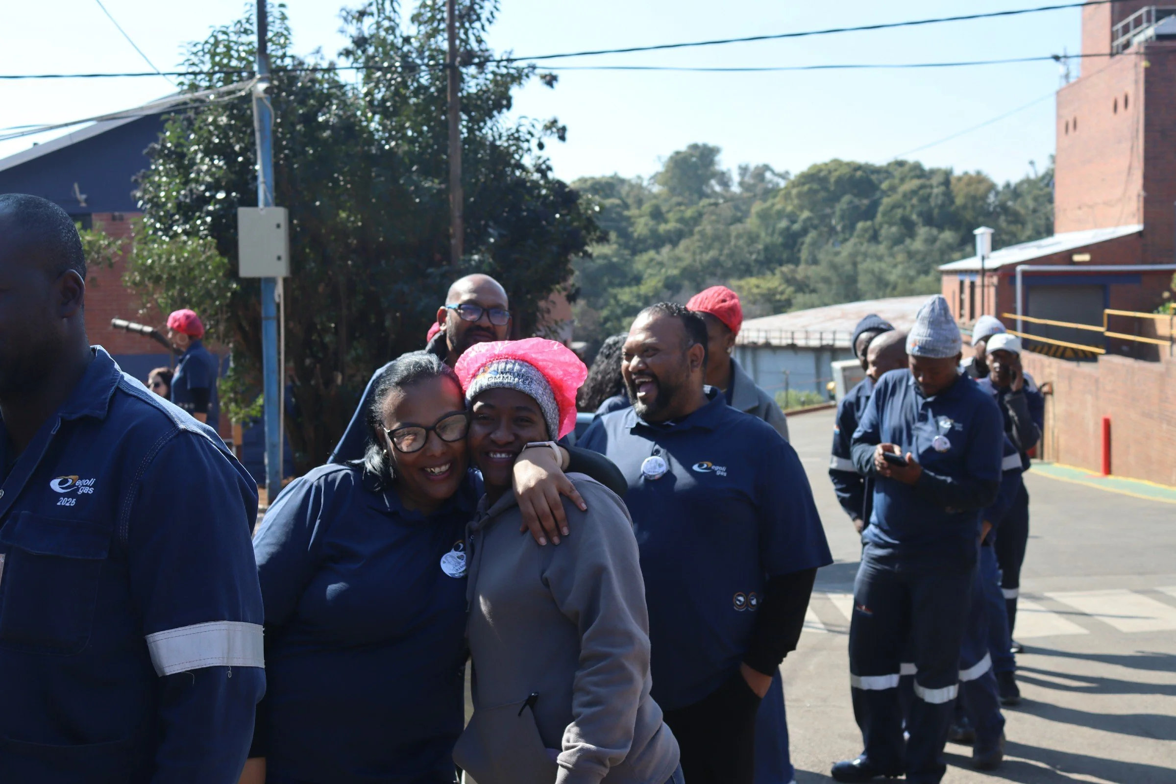 A group of smiling people in blue uniforms gather outside on a sunny day, some embracing and others looking at their phones, with trees, buildings, and power lines in the background.