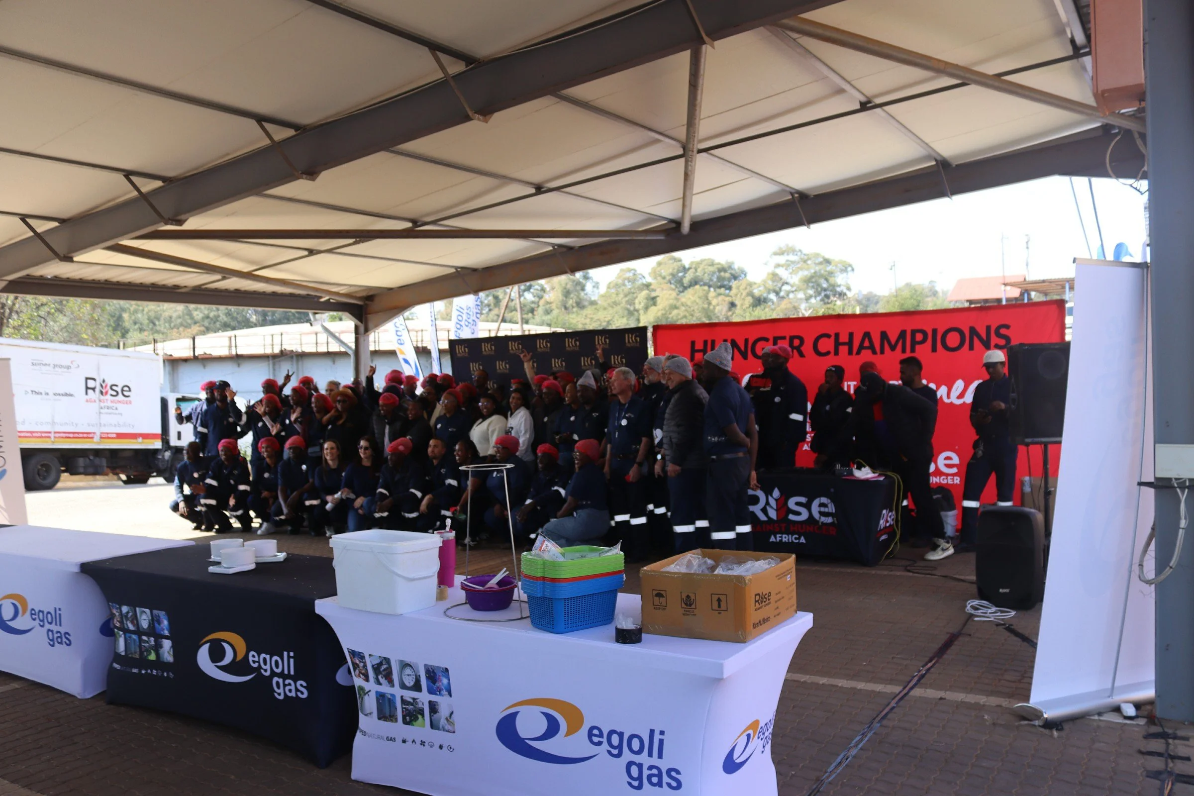 A large group of people gathered at an outdoor event under a tent, with some participants wearing red hats. The event appears to be related to Hunger Champions, as indicated by the large red banner. There are tables with items, including cans and box