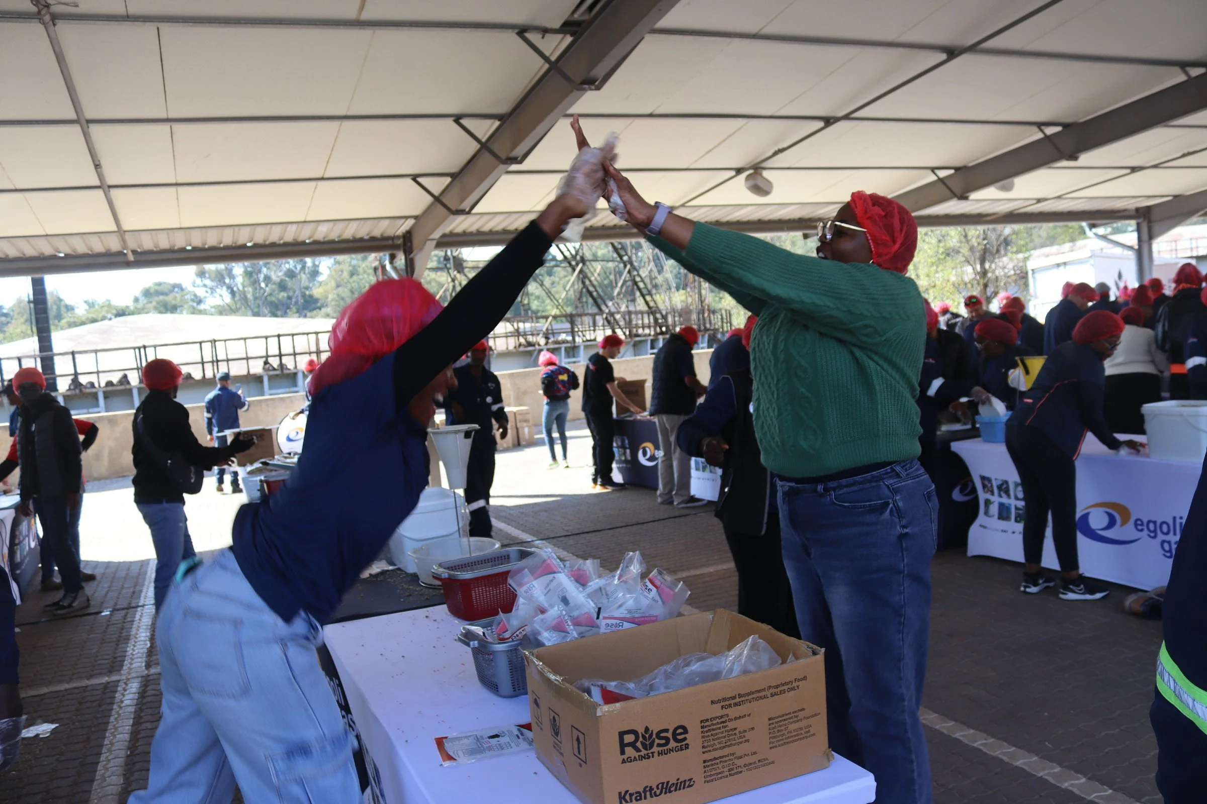 People participating in a community event, handing out food and supplies under a large covered outdoor area. Many are wearing red hairnets and dark clothing, with some reaching out for high-fives. There are tables with boxes of food and supplies, and