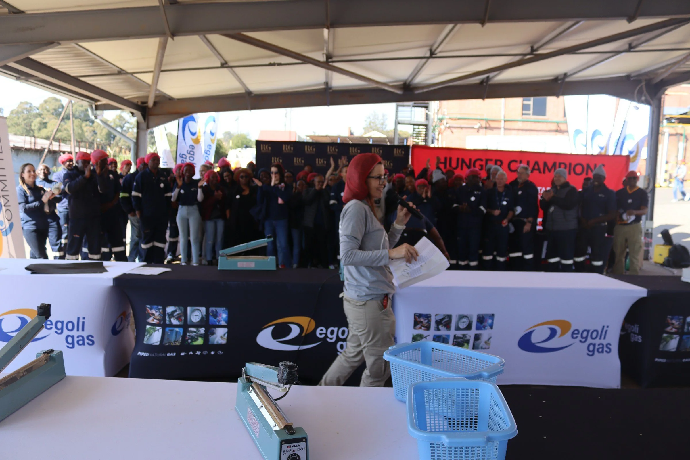A woman speaking into a microphone during an outdoor event, with many people in uniforms and red hats standing behind her under a large tent. There is a red banner that says 'HUNGER CHAMPION' and a black banner with golden letters.