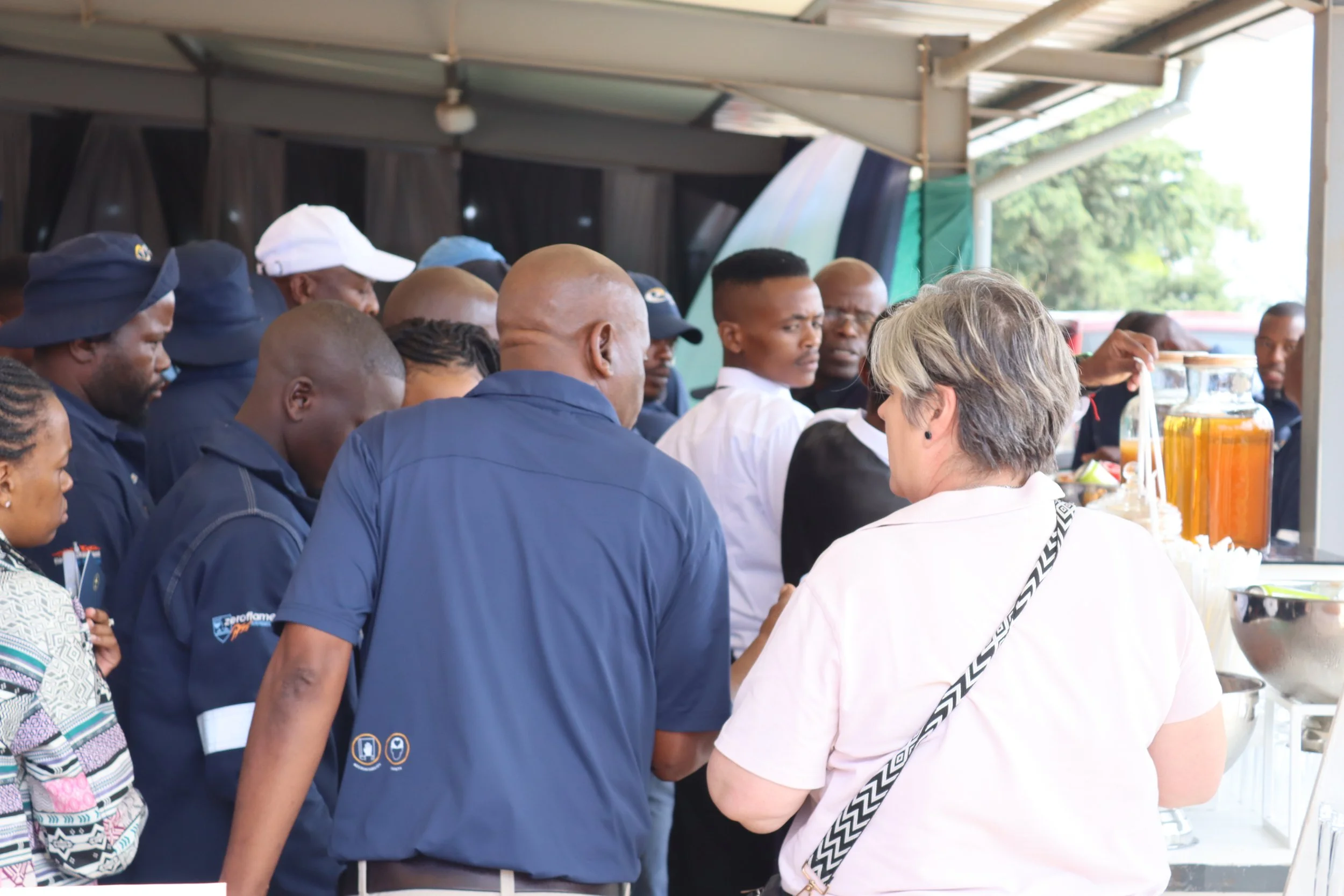 A group of people gathered at a food stand under a metal roof, with some holding money and others waiting to order. The stand offers beverages in containers, possibly juices or iced drinks.