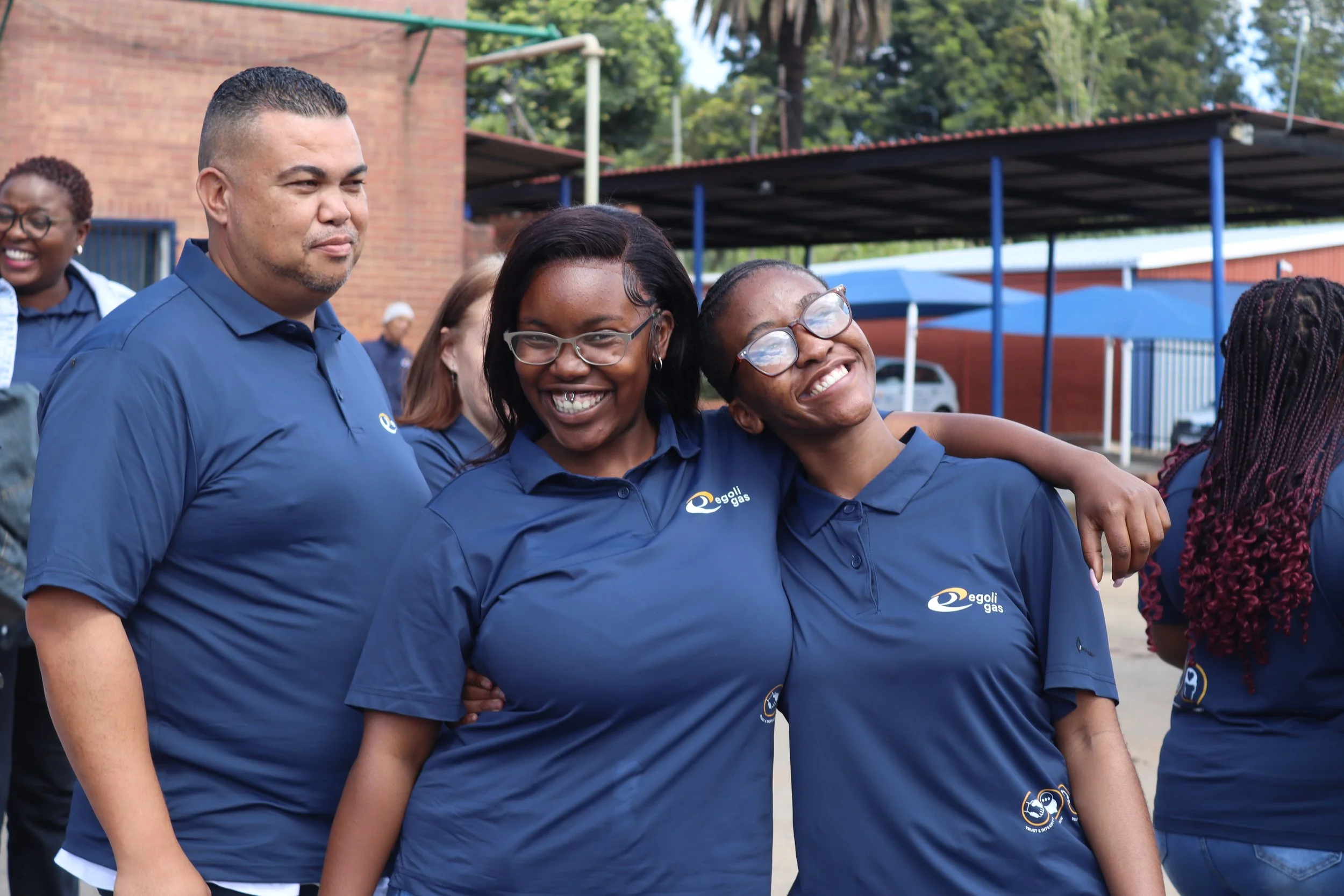 Group of smiling people outdoors wearing blue shirts, some with glasses, standing close together with arms around each other.