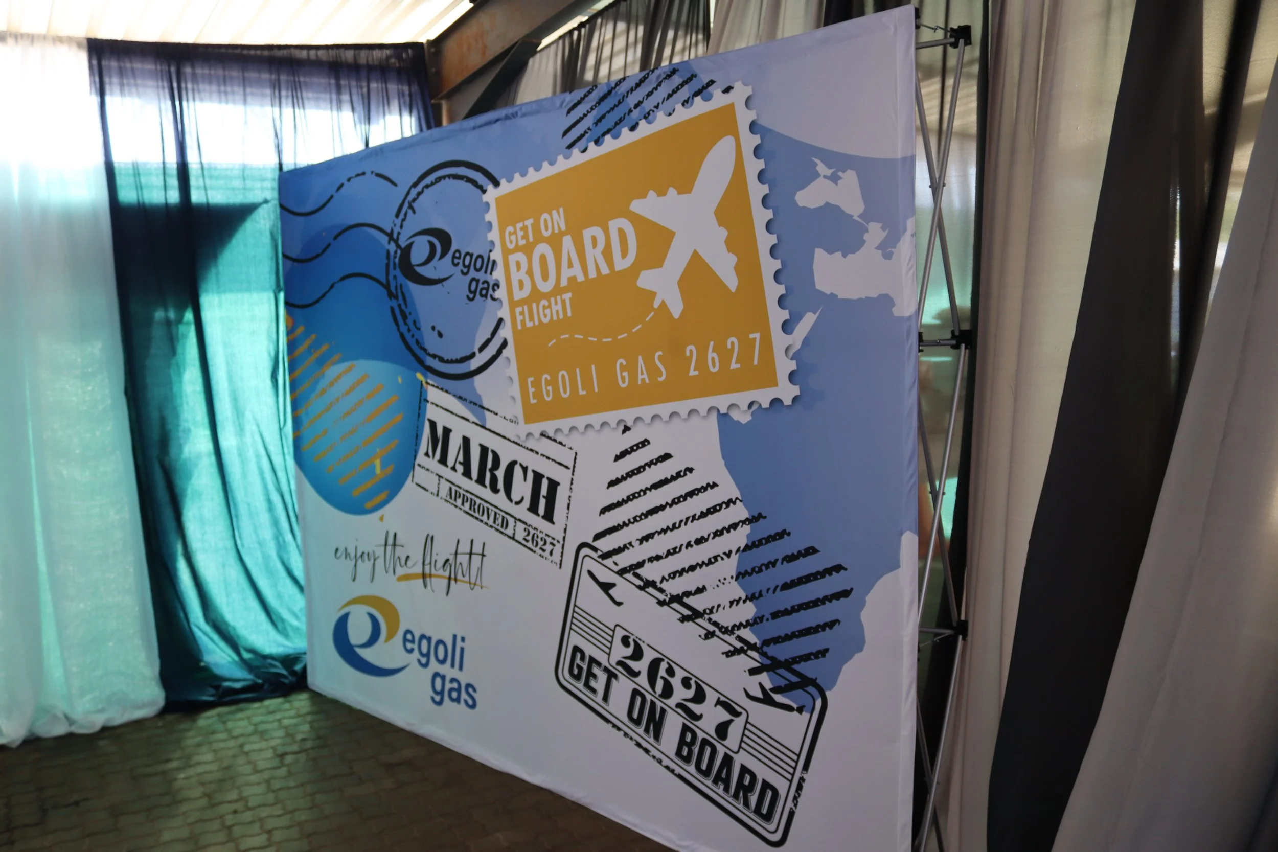A large sign for the Egoli Gas company featuring a yellow airplane graphic and text that says "Get on Board Flight" and "Egoli Gas 2627." The sign also includes a black and white stamp design with the message "Get on Board" and the date March 2023.