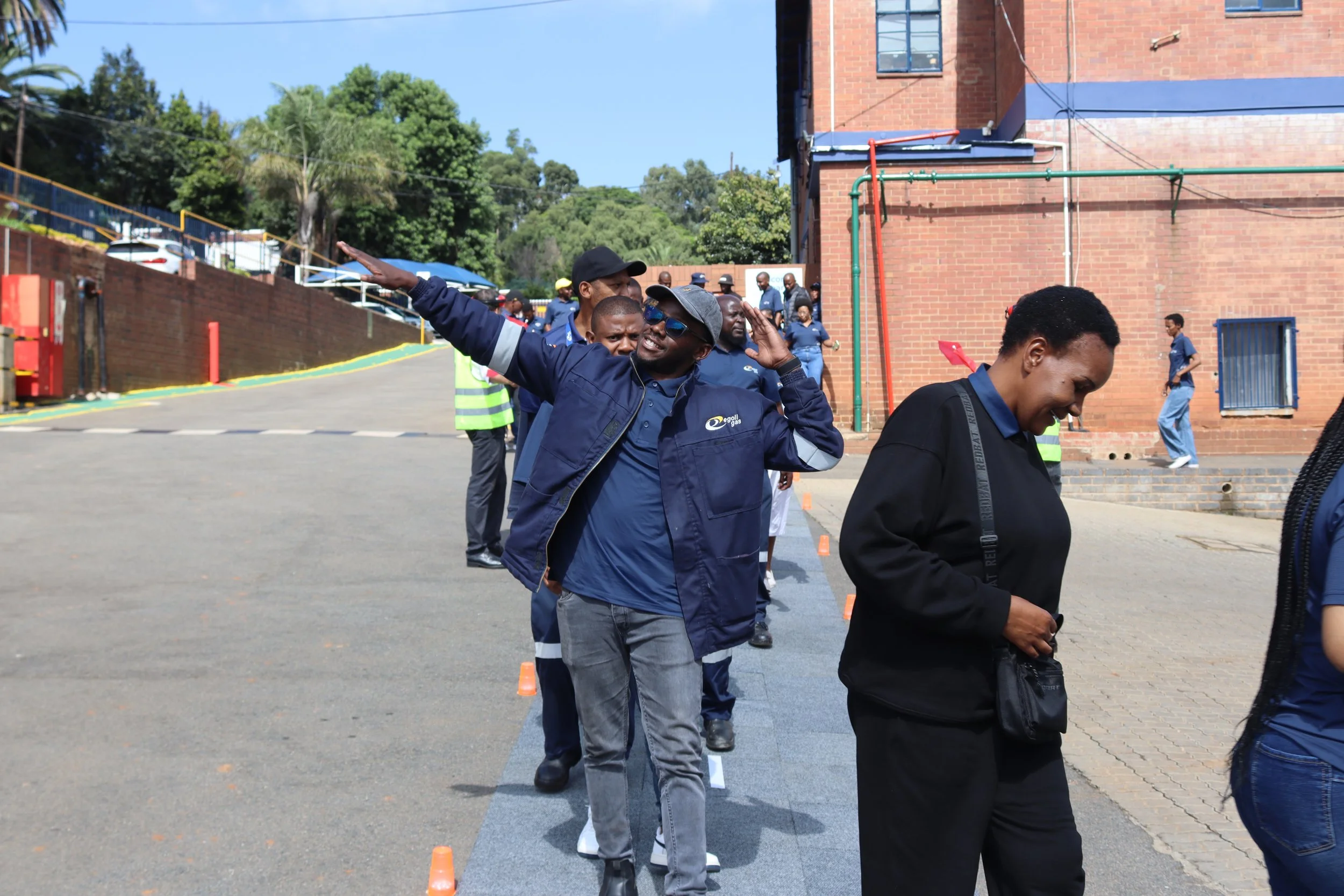 A group of people standing outdoors in a line, participating in an organized activity or event on a sunny day, with a brick building and green trees in the background.