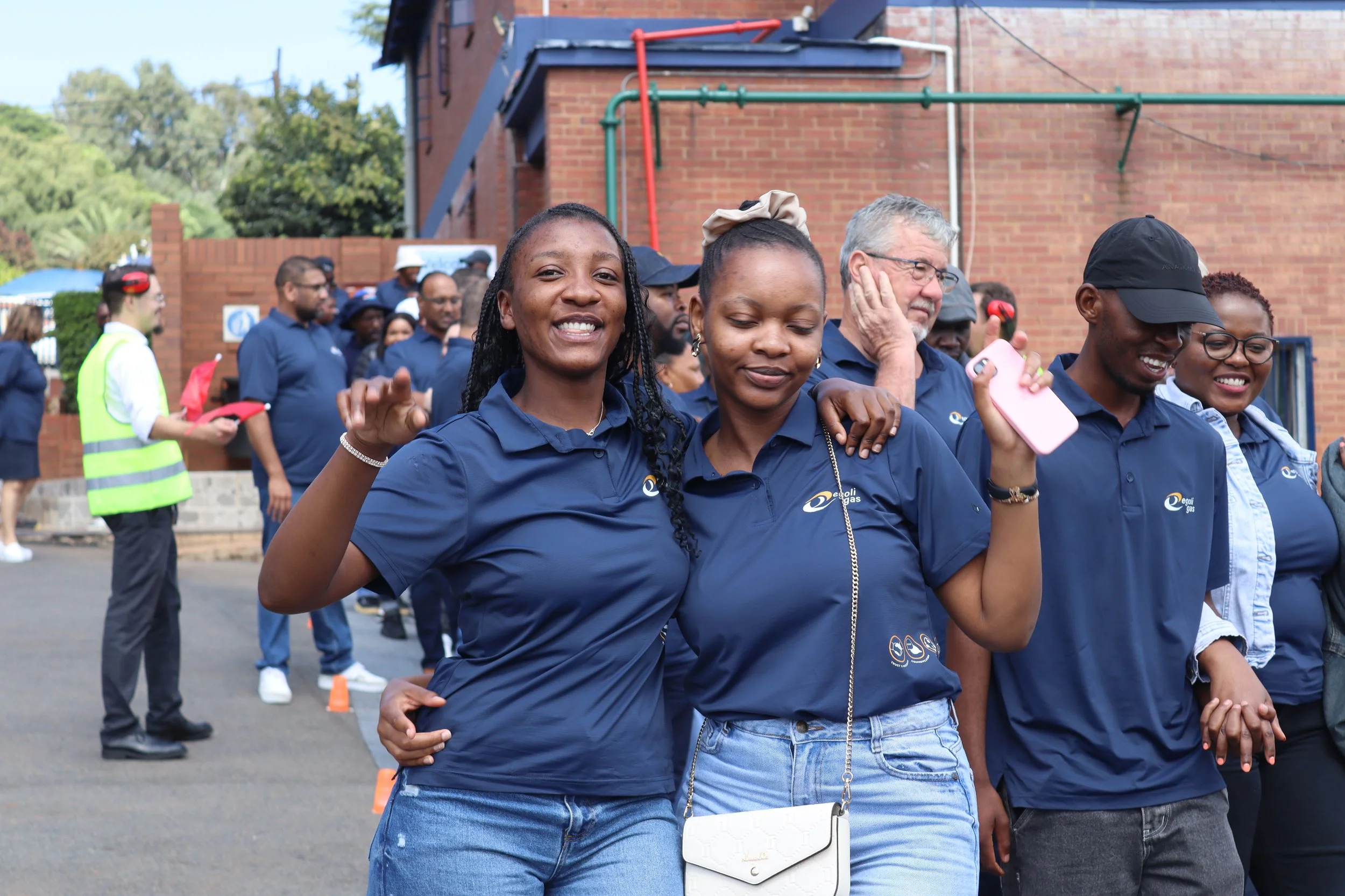 Group of diverse people at an outdoor event, some smiling, some looking contemplative, with a brick building and trees in the background.