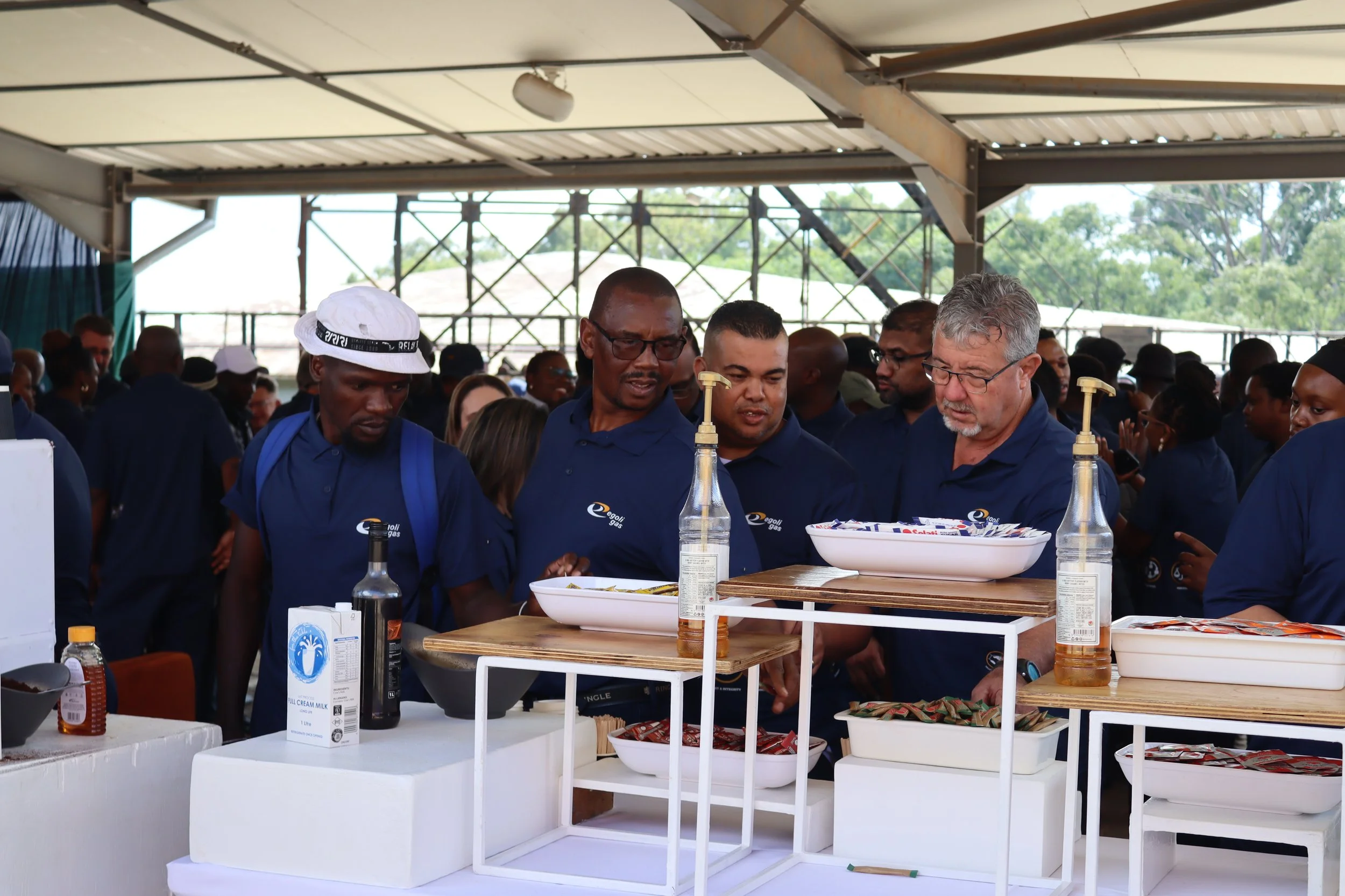 Several people gathered at an outdoor event, wearing blue shirts with a logo, standing around a table with food and drinks. The background shows a large open space with a metal framework and trees.