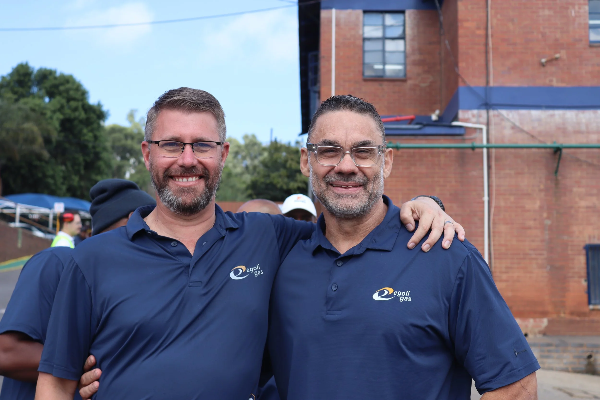 Two men smiling and standing close together outdoors, wearing navy blue shirts with the logo 'Egoli Gas' on them. One man has glasses and a beard, the other has short hair and a beard. They are embracing each other with their arms around each other's