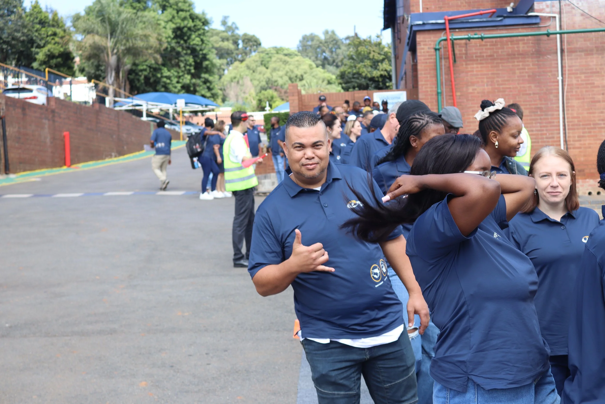 A group of diverse people wearing matching navy blue shirts, gathered outdoors near a brick building, some smiling and giving a thumbs-up. The scene appears to be at a community event or gathering.