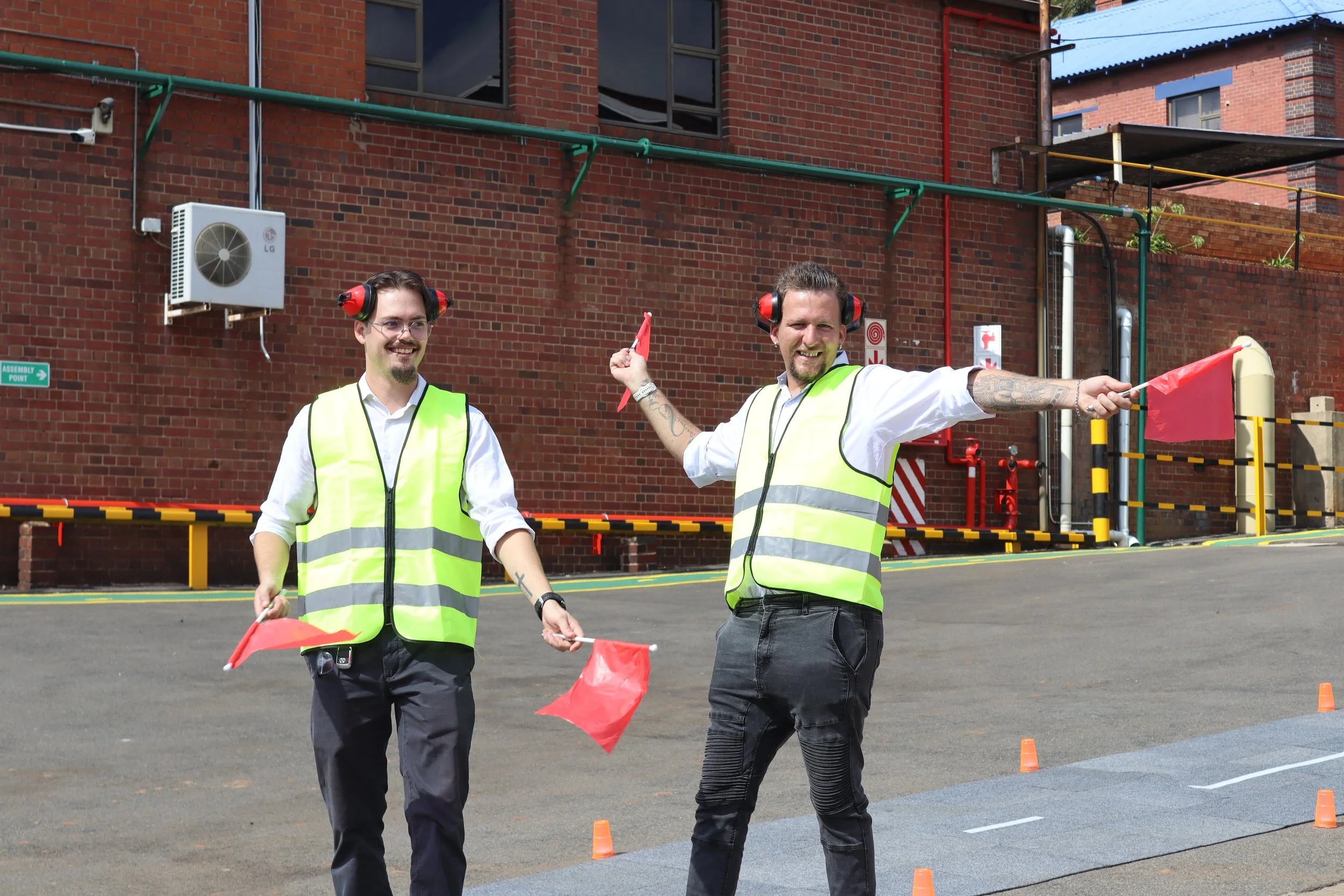 Two men wearing yellow safety vests and red ear protection, holding red flags, participating in an outdoor event with orange traffic cones in front of a brick building.