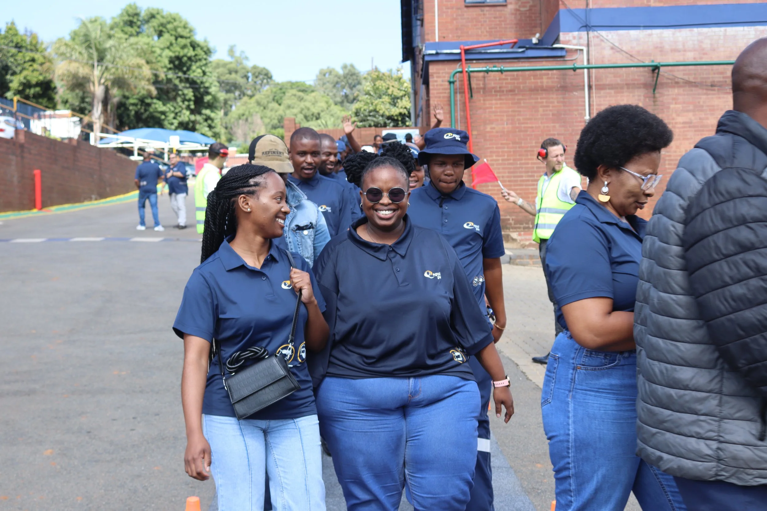 Group of smiling women and men walking outdoors, wearing dark blue shirts with a logo, some wearing hats and sunglasses, in front of a brick building and trees.