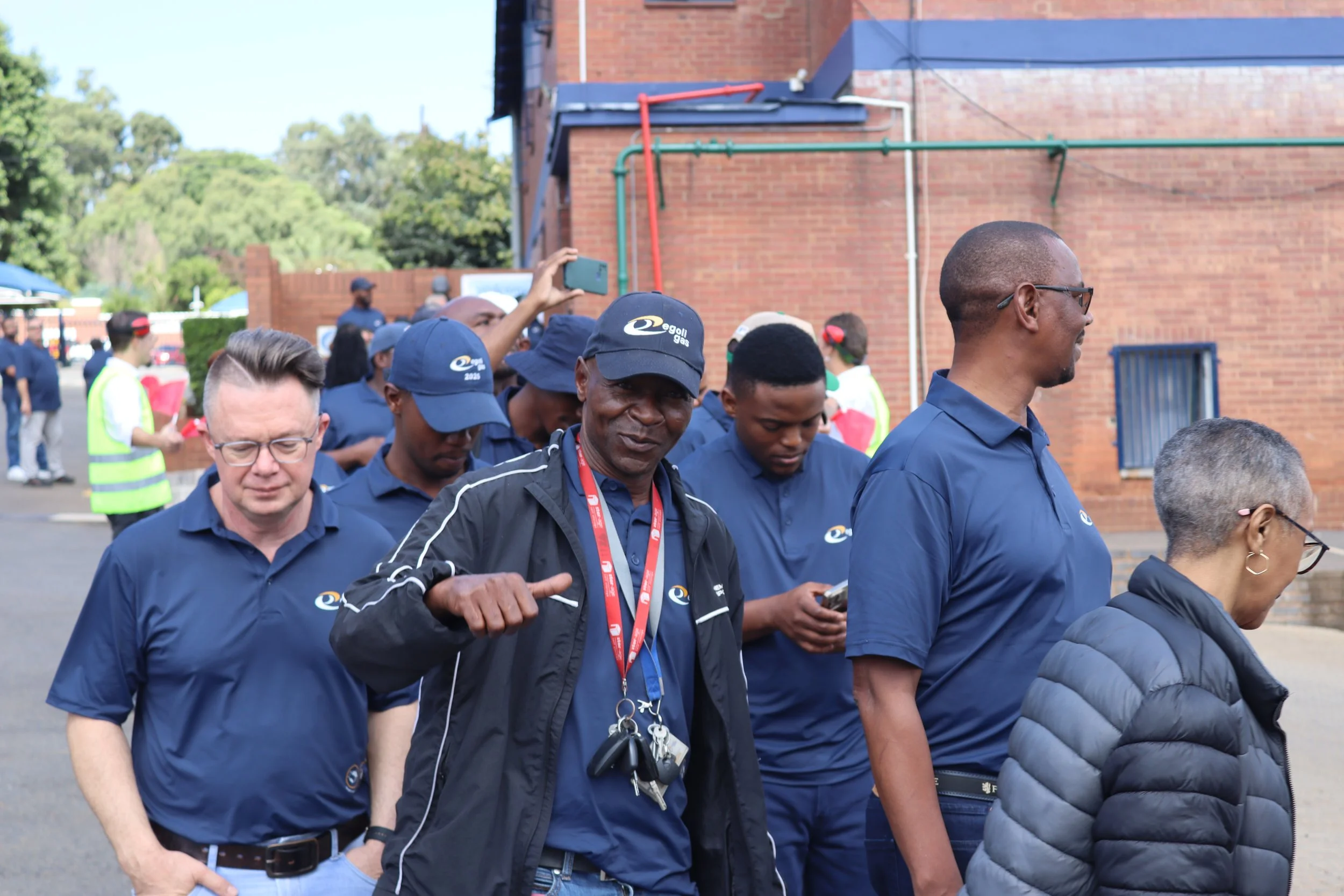 A group of people, mostly men, wearing navy blue shirts and hats, walking outdoors near a brick building. Some are looking at their phones or talking. One person is holding a camera or phone up to take a picture.