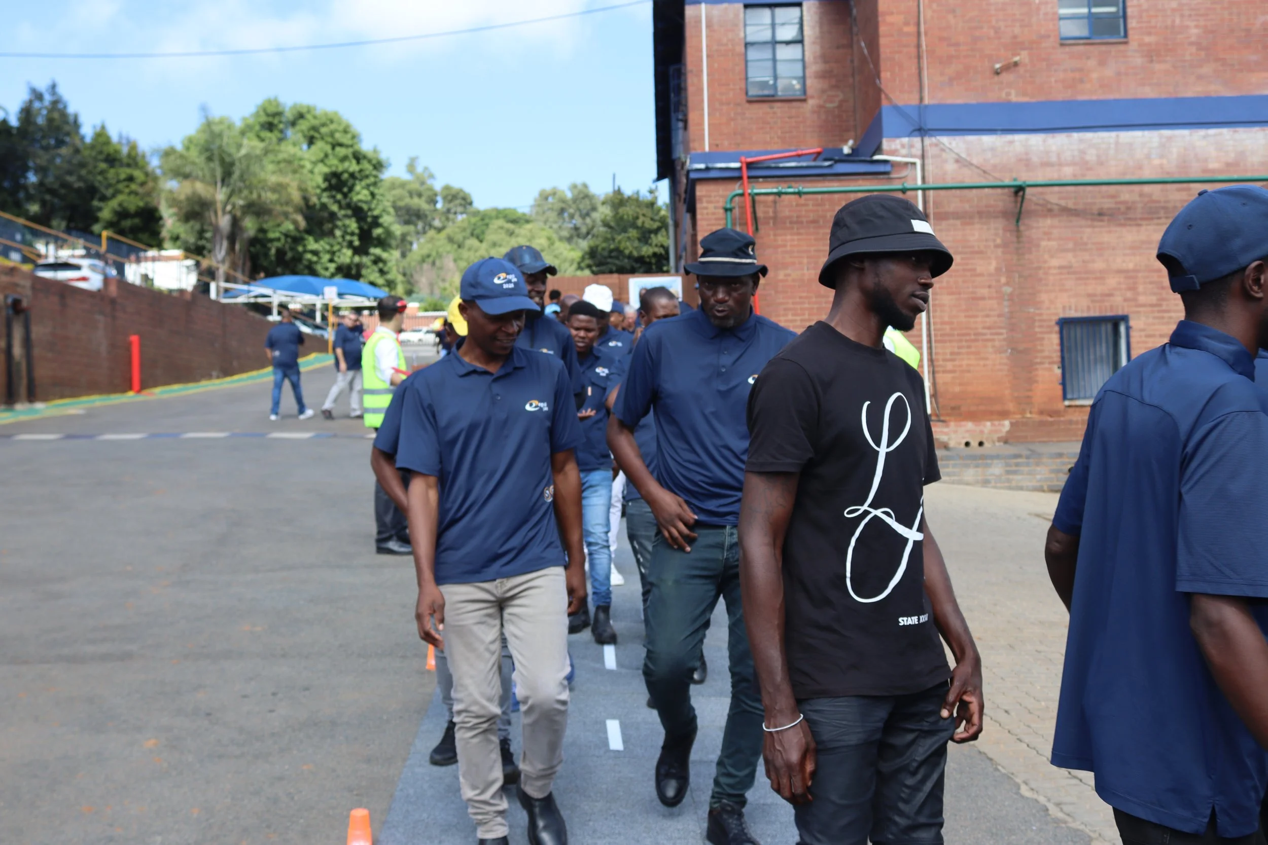 Group of people participating in an outdoor event, walking in a line behind orange cones on a paved area, with a brick building and trees in the background.
