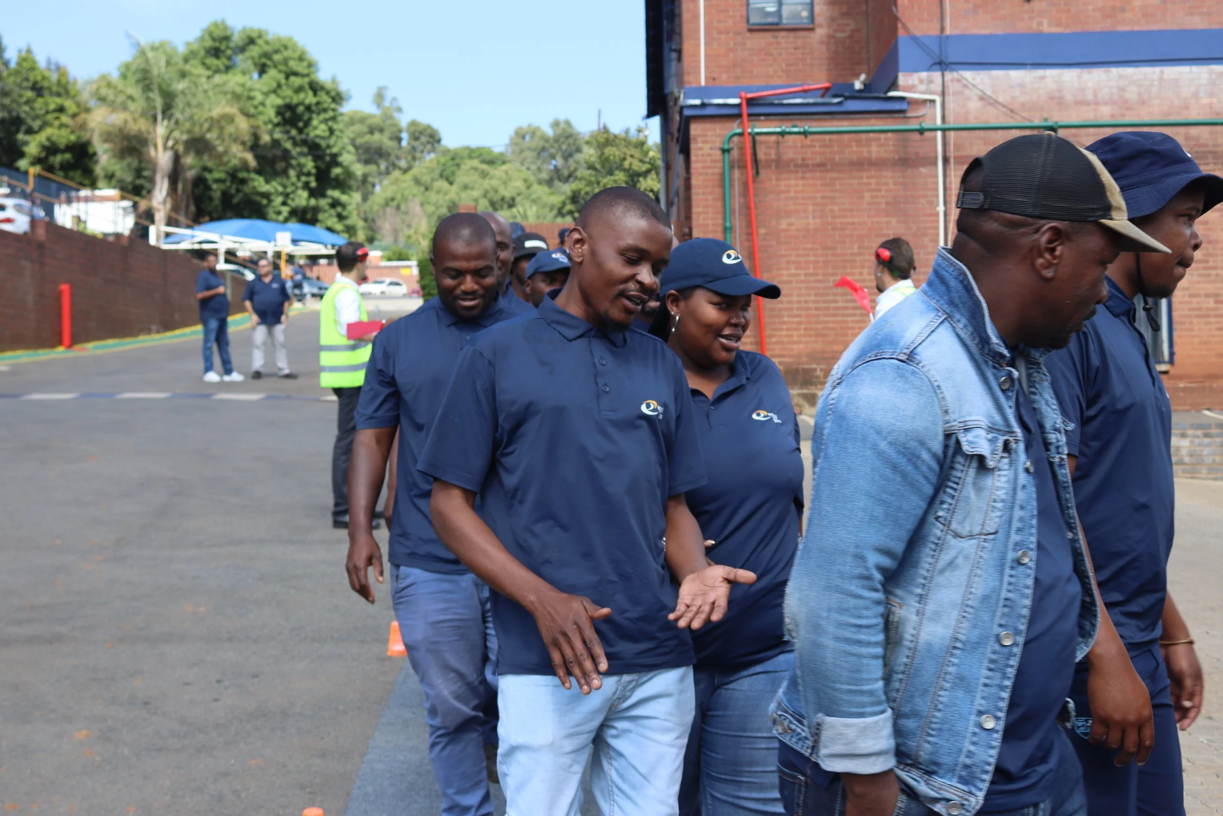 A group of people walking in a line outdoors, with several individuals wearing blue shirts and a woman wearing a blue cap. In the background, there are trees, a brick building, and a few other individuals standing or walking near orange cones.