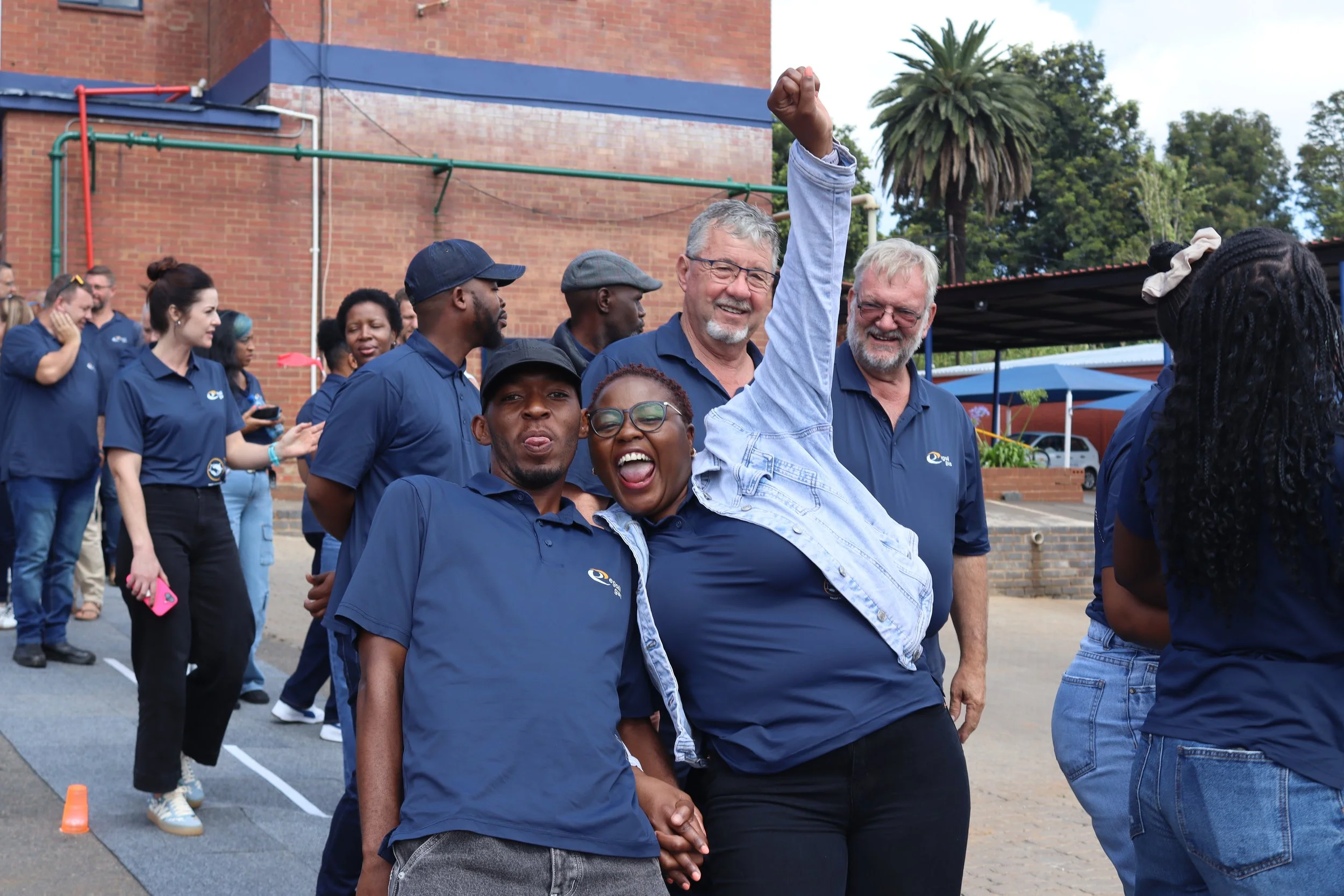 Group of diverse people wearing blue shirts outdoors, celebrating and holding hands, with a brick building and trees in the background.