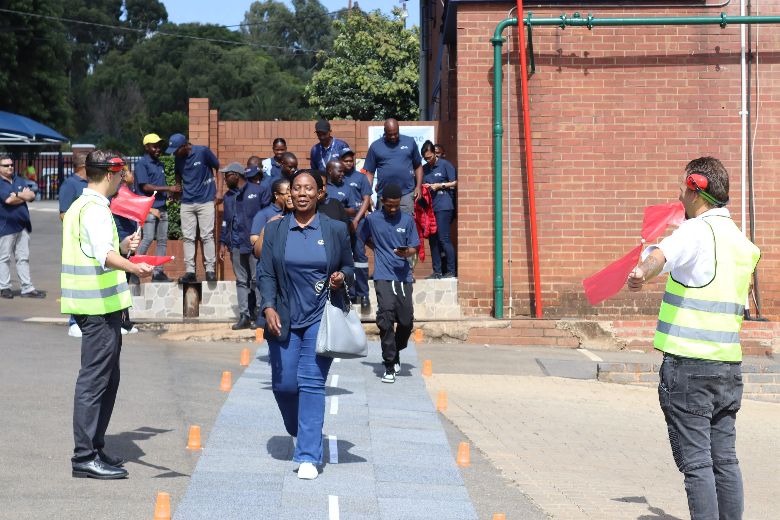 A group of people, including men, women, and children, participating in an outdoor event near a brick building under a sunny sky, with some individuals holding small flags, and others walking or standing around.