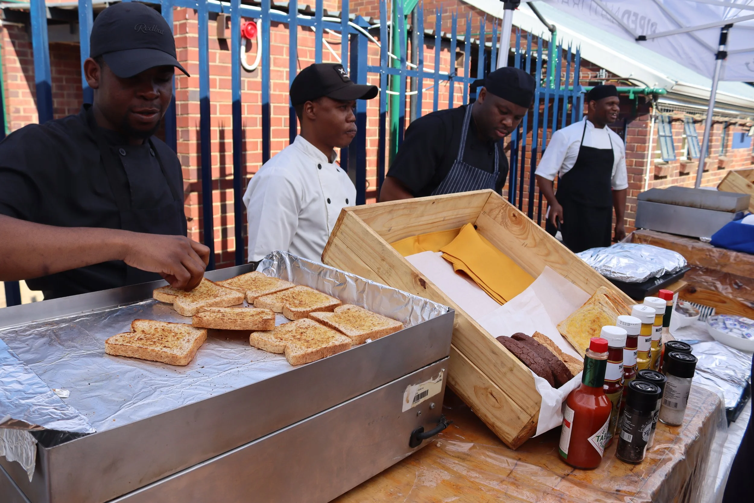 Four chefs preparing food at an outdoor event stand, with bread, condiments, and grilled items on display, against a backdrop of brick wall and blue metal fence.