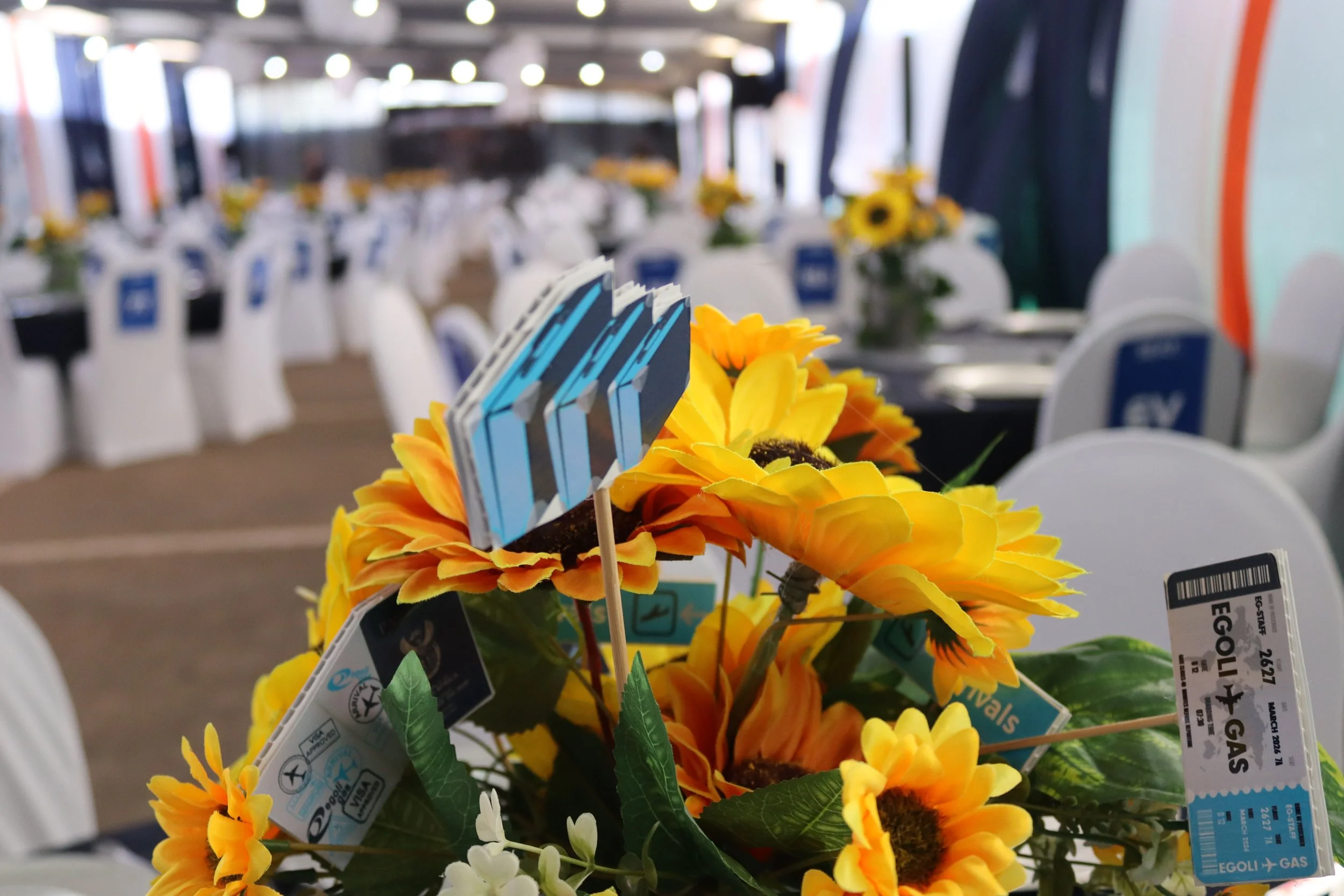 Close-up of artificial yellow sunflowers with travel destination tags on plants, in an airport waiting area with seats and blurred background lights.