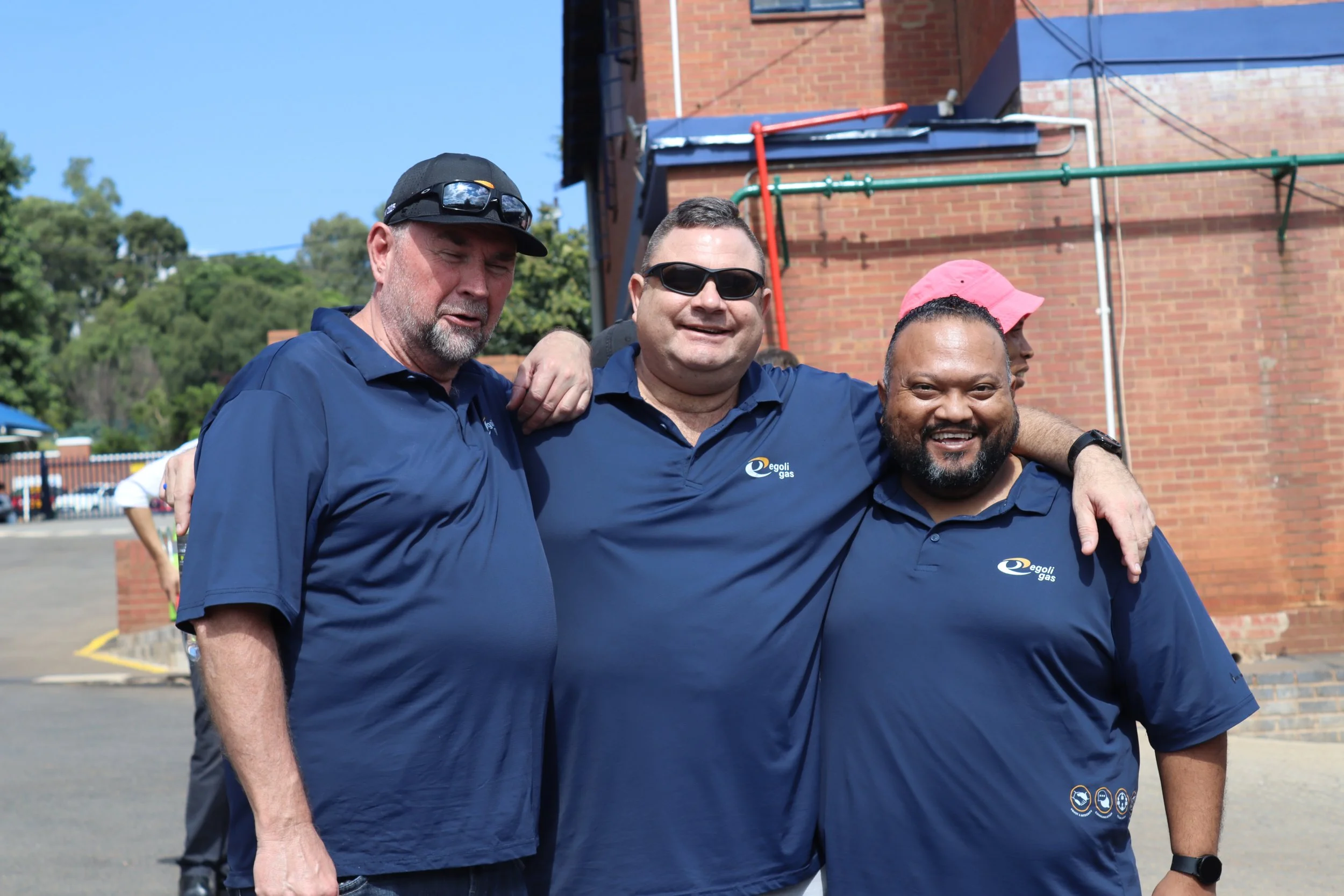 Three men smiling, wearing blue shirts with a logo, standing outdoors with a brick building behind them. One man is wearing sunglasses, and another has a pink hat. The middle man has his arm around the others.