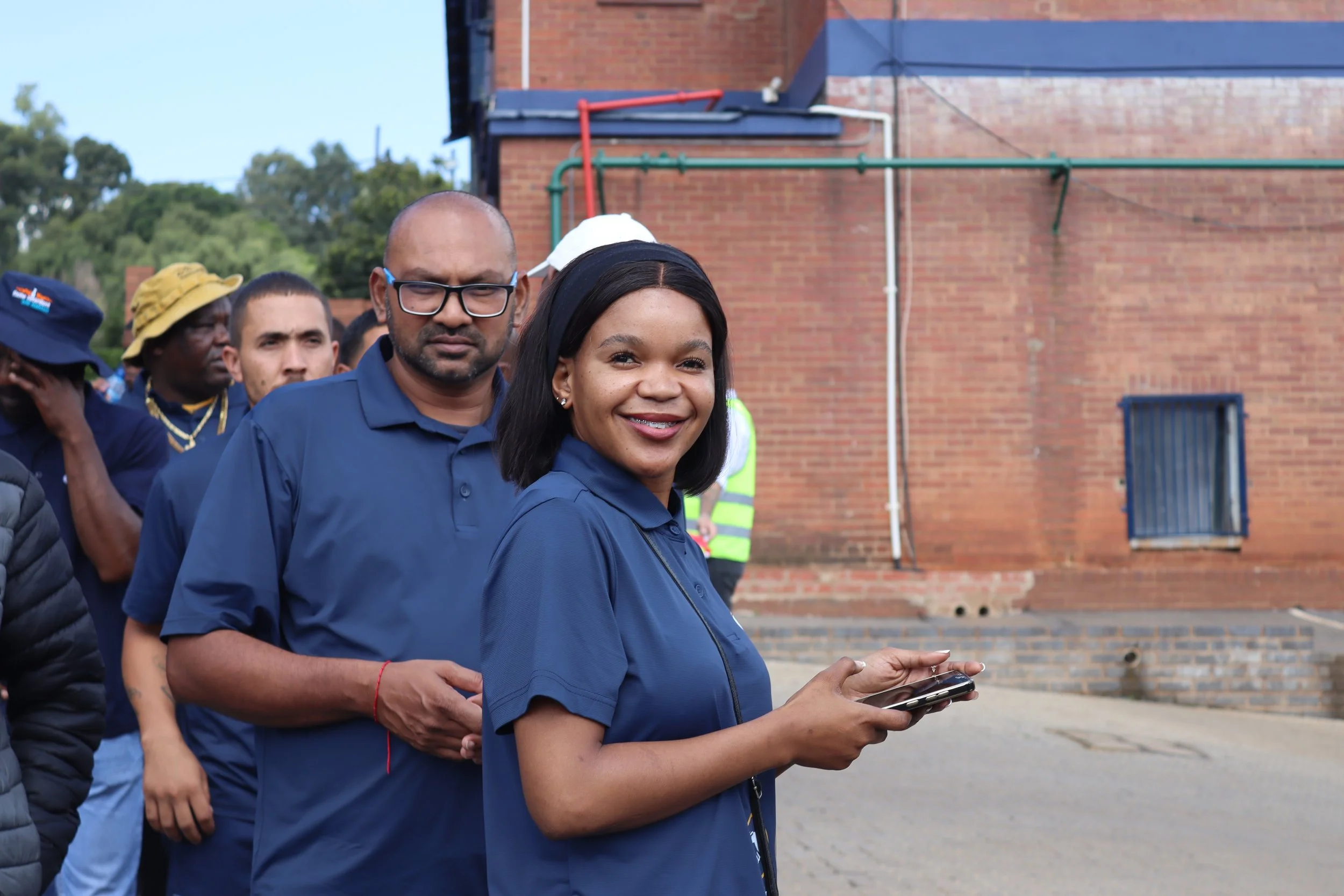 A group of people standing in line outdoors, with a woman in front smiling and holding a phone, and a man behind her looking at the camera. The background features a brick building and some greenery.