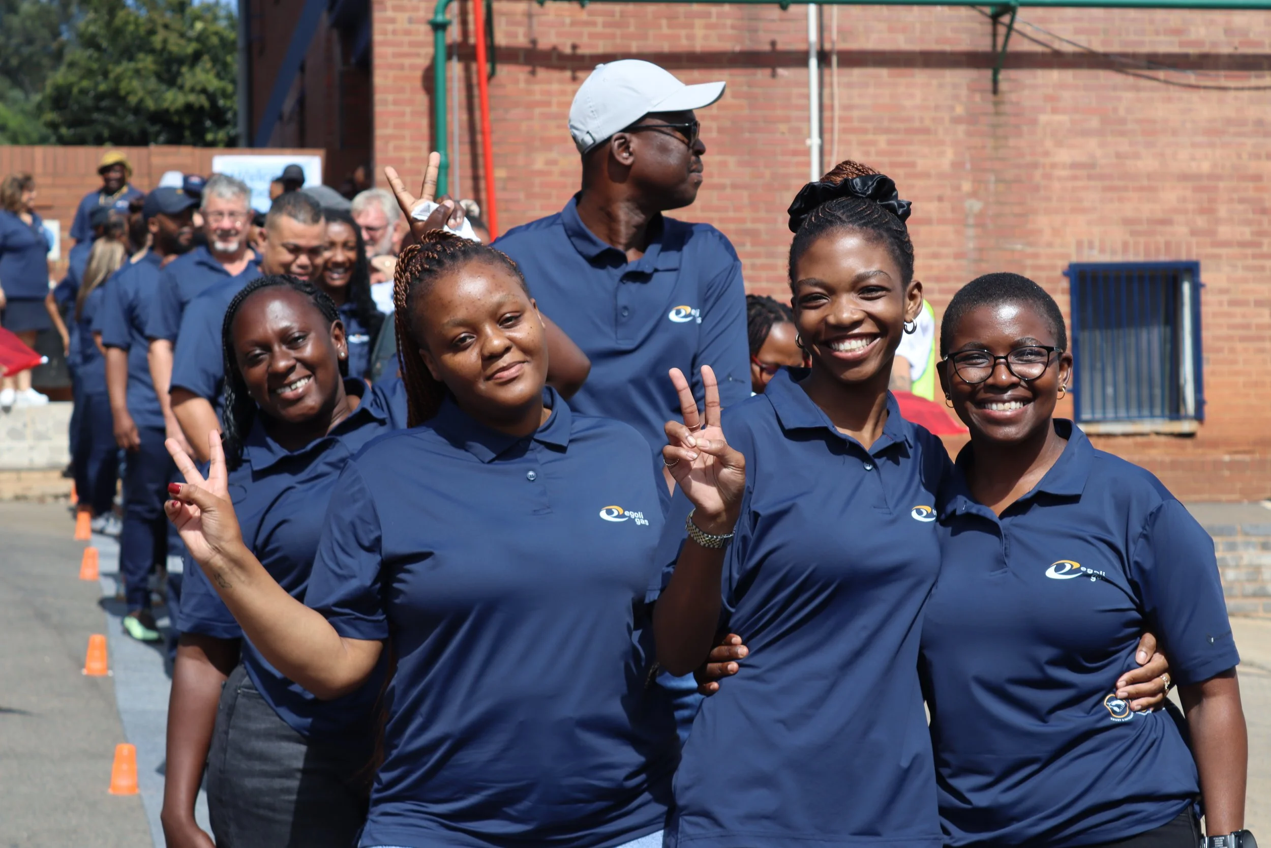 Group of people standing in a line outdoors, wearing matching navy blue shirts, some making peace signs, with a brick building in the background.