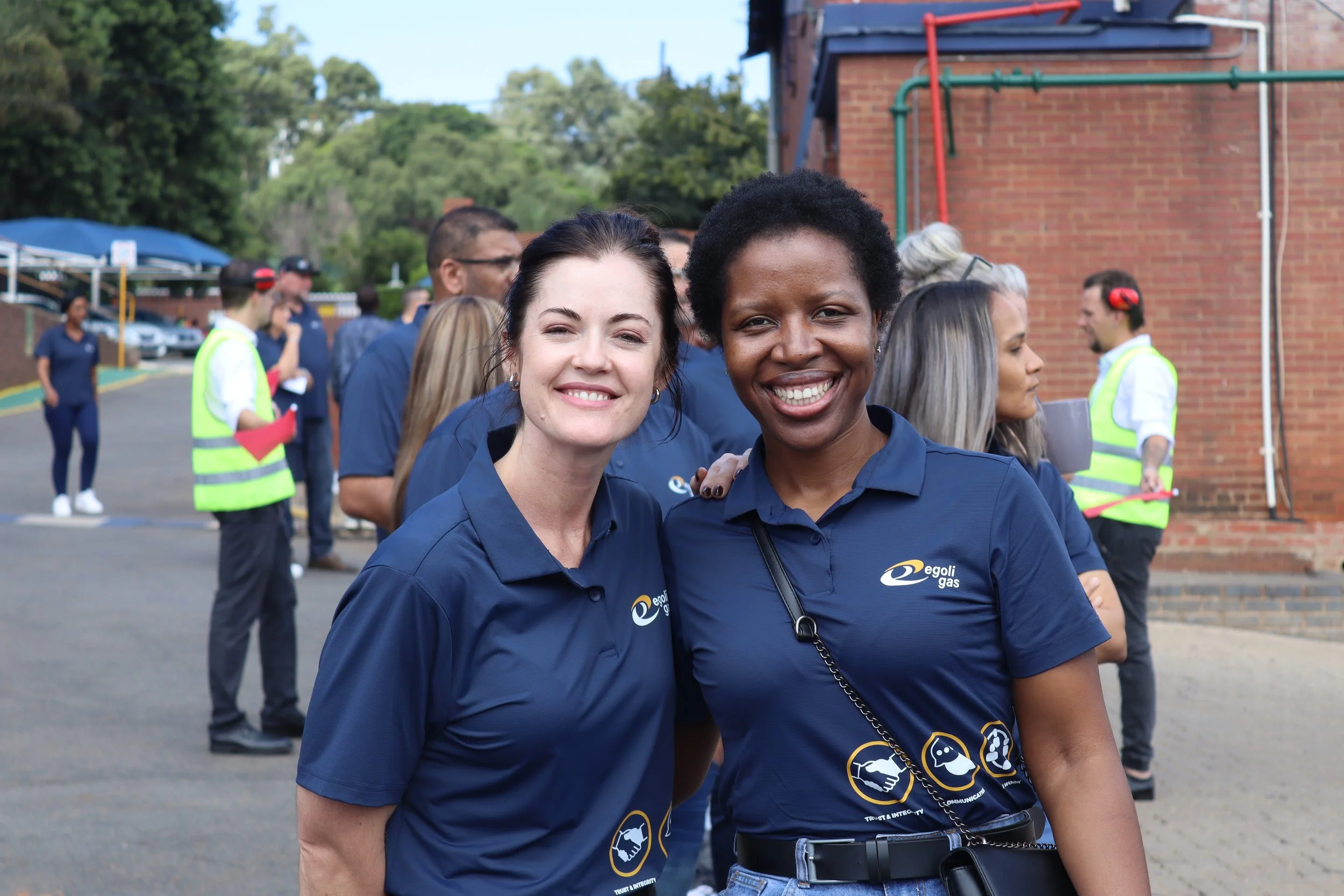 Two women smiling at the camera, wearing navy blue shirts with a logo, standing outdoors with a group of people in the background.