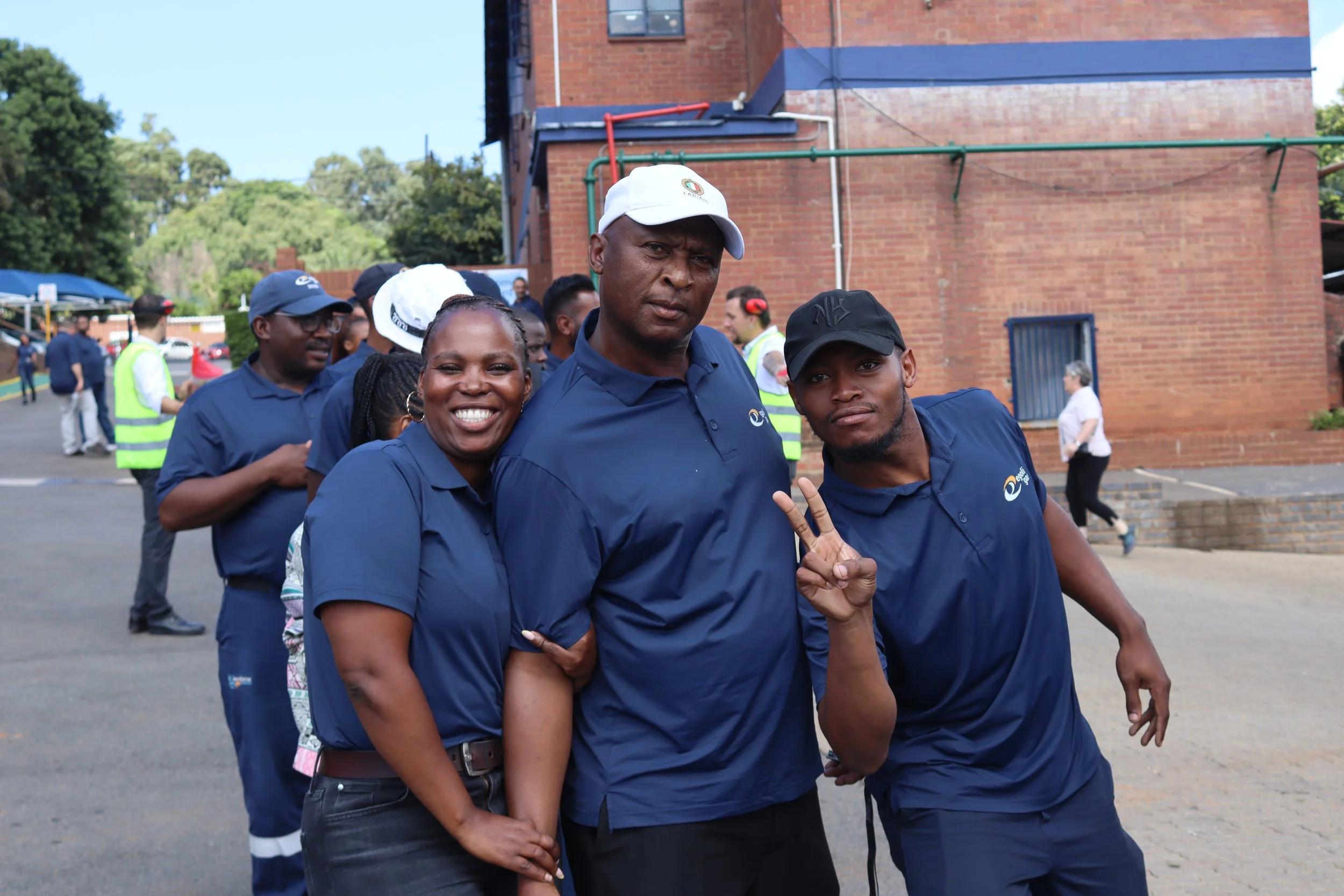 Group of people wearing dark blue shirts outdoors, some smiling and making peace signs, with brick building and trees in the background.