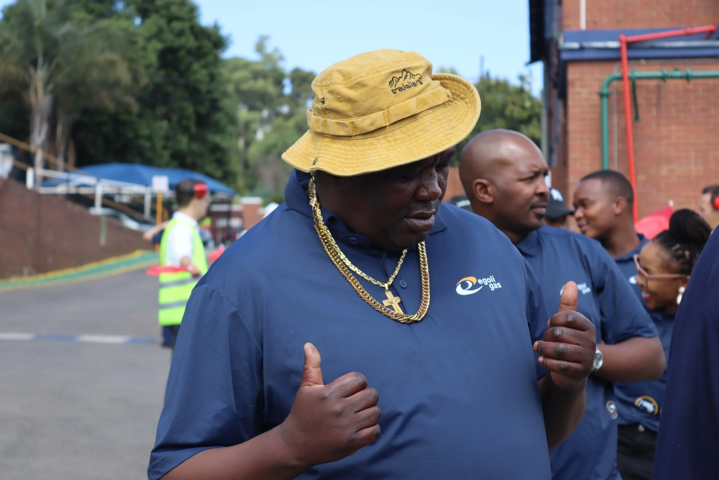 A man wearing a yellow bucket hat, gold chains, and a blue shirt with a logo interacts with another person in a group setting outdoors, with trees and a brick building in the background.