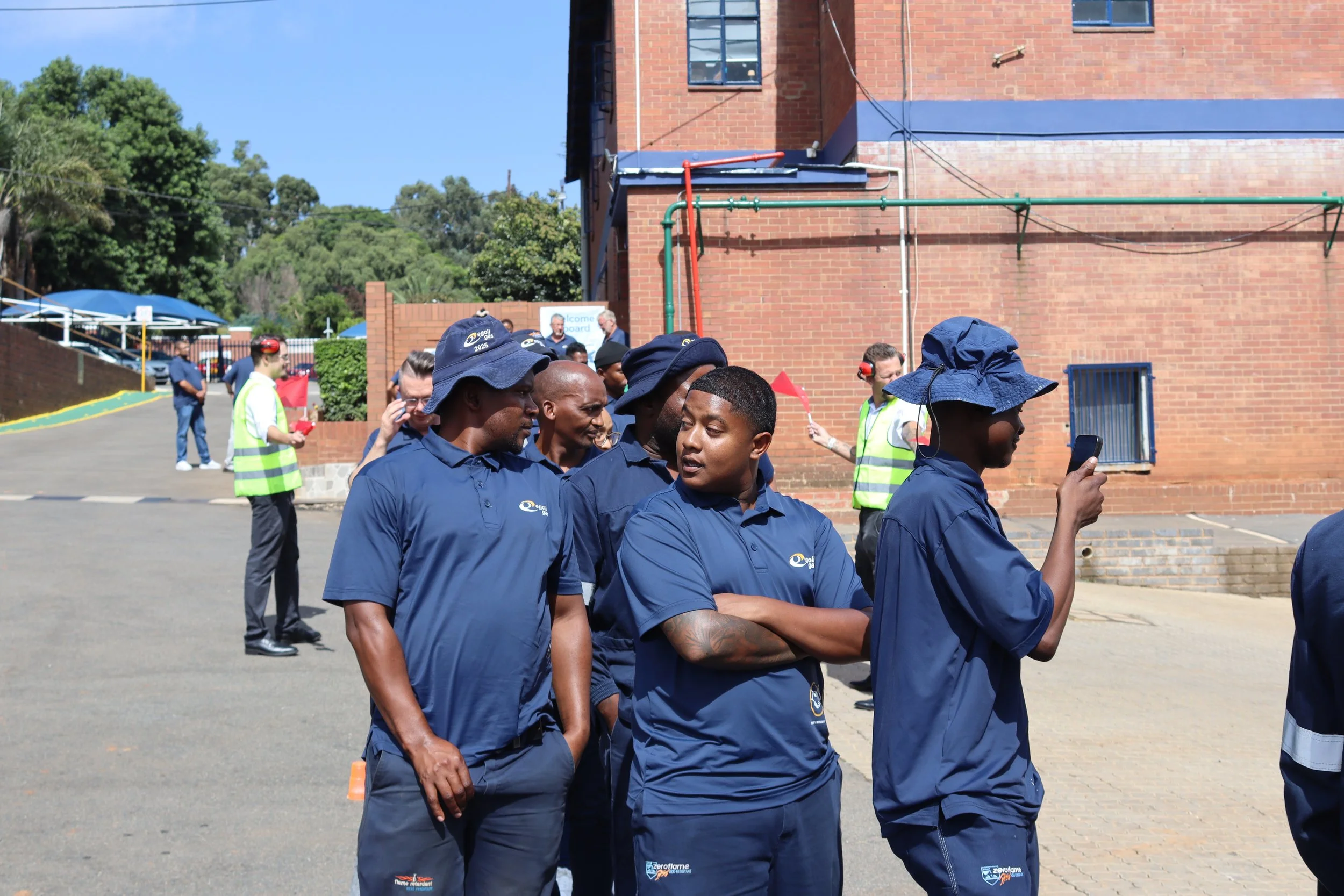 A group of young men in navy blue uniforms standing outdoors in front of a brick building, some wearing hats and one using a smartphone. Several people in the background are supervising or observing, with some in casual attire and reflective vests, u