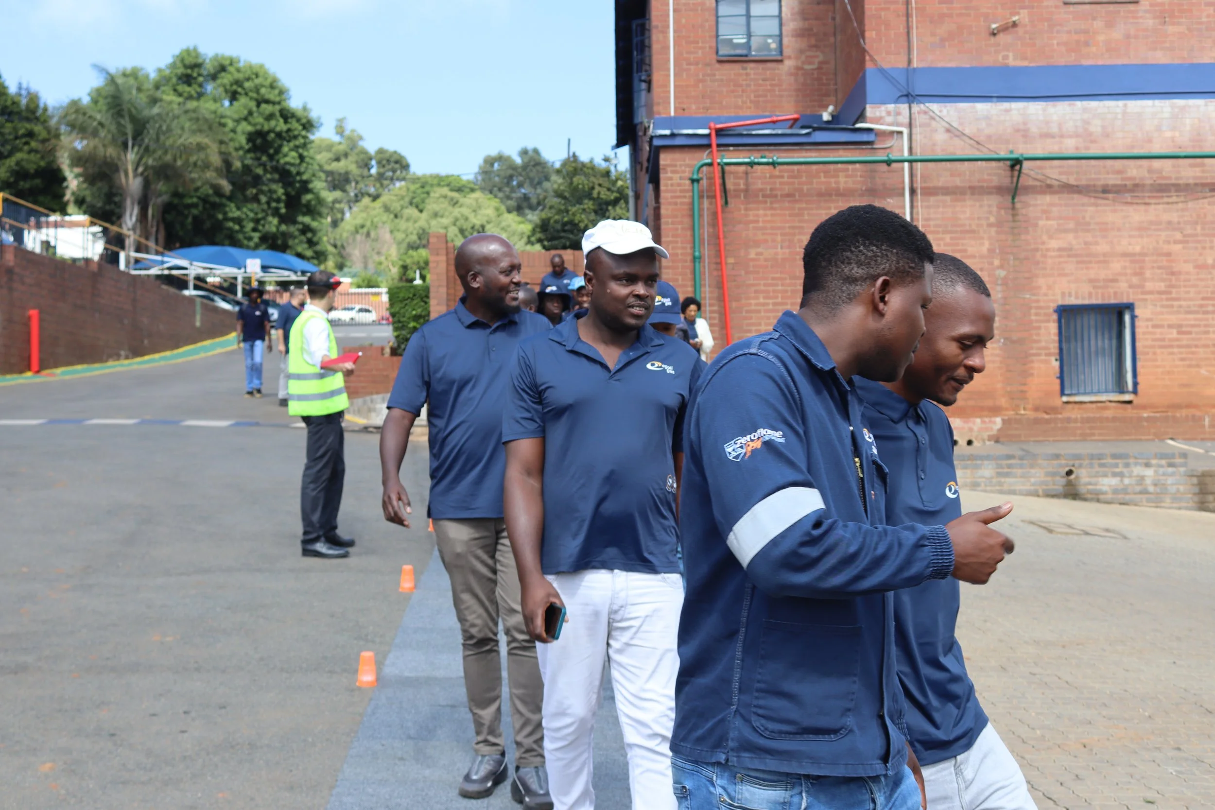 Group of men standing in line outdoors, some looking at their phones, in front of a brick building with trees and blue sky in the background.