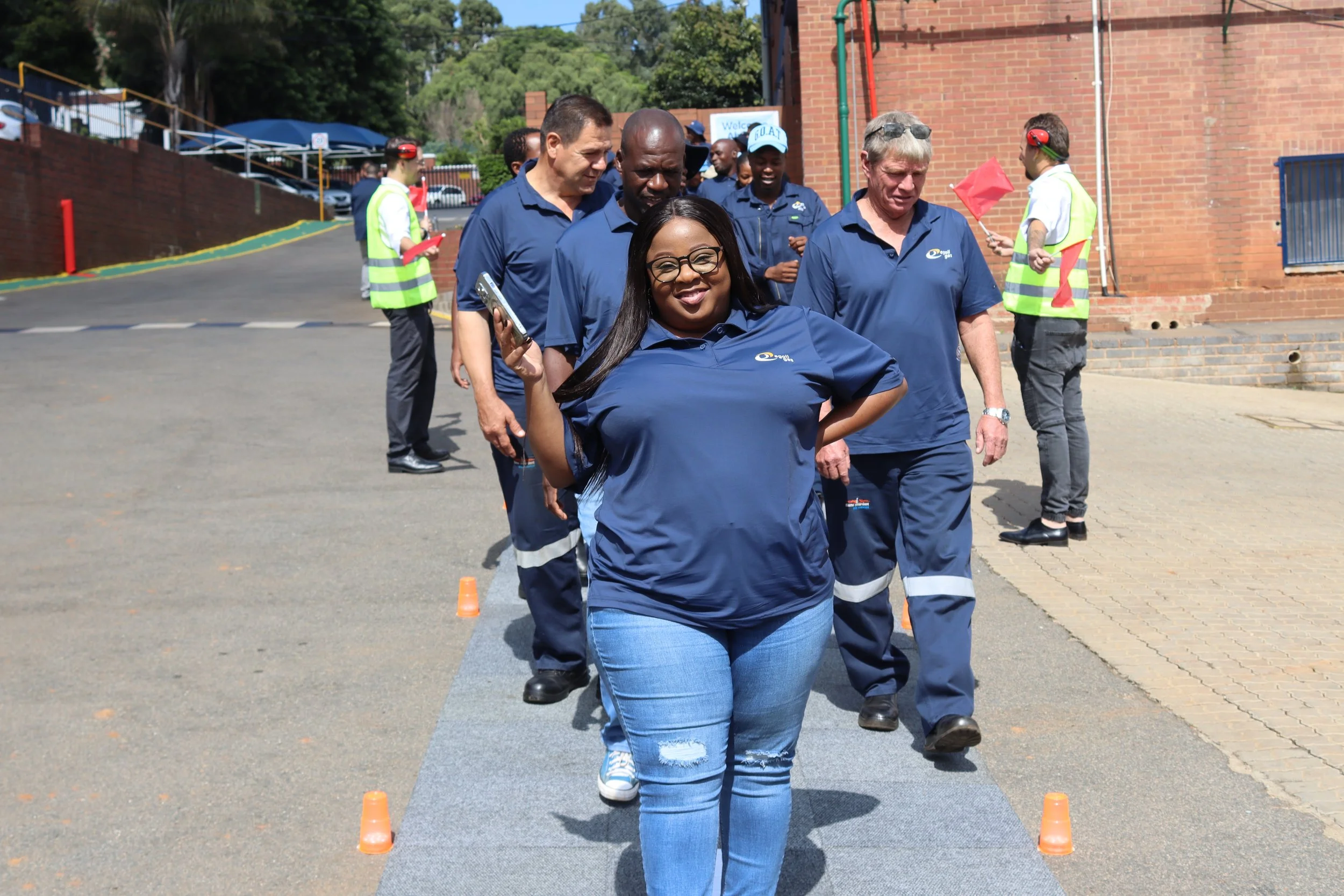 Group of diverse people participating in an outdoor team-building activity involving walking through orange cones on a paved surface, with some holding clipboards and others wearing safety vests.
