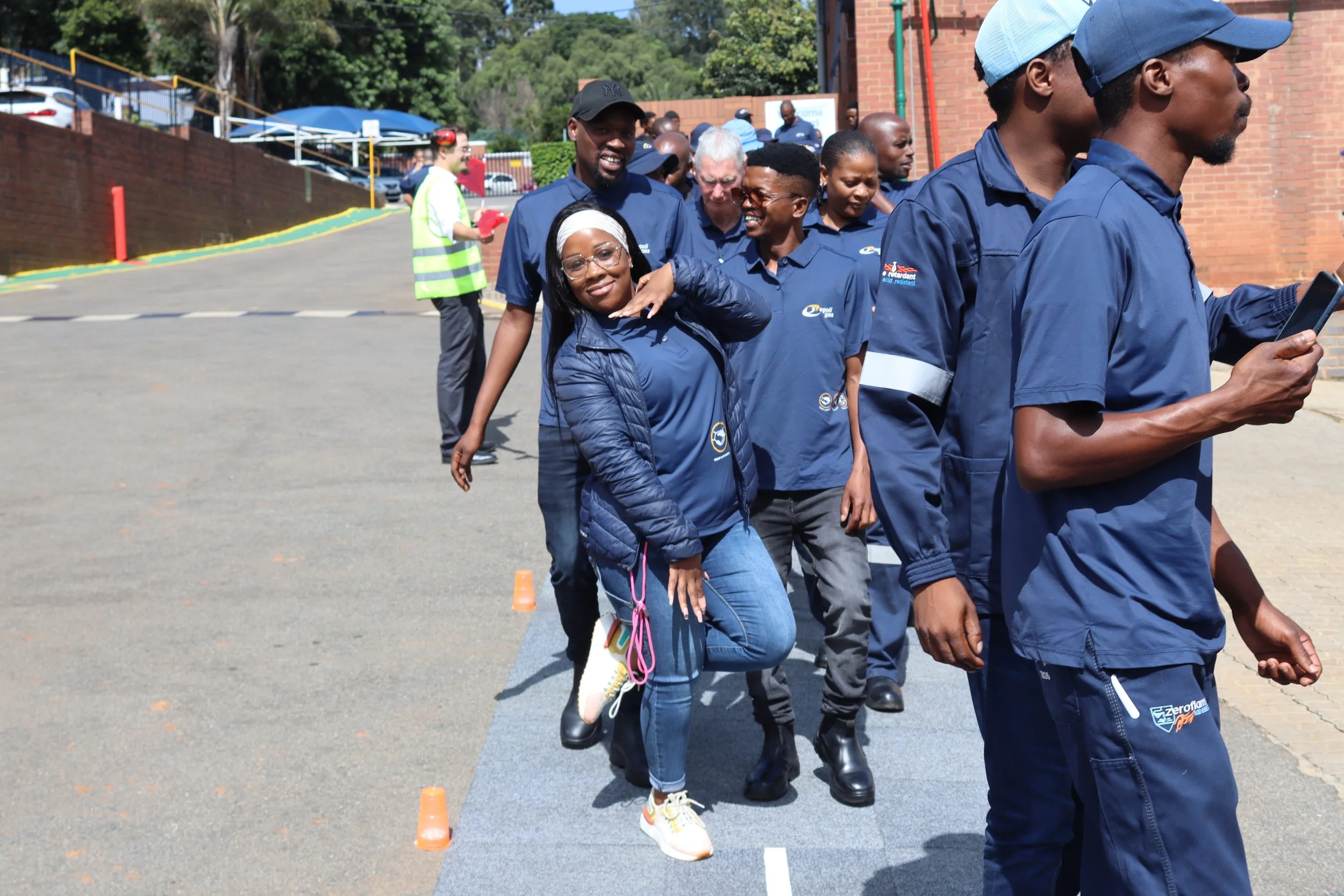 A group of people participating in an outdoor team activity involving rope. They are standing in a line with some smiling and others concentrating, with cones set up on the ground. All are dressed in blue uniforms, and there is a woman in the foregro