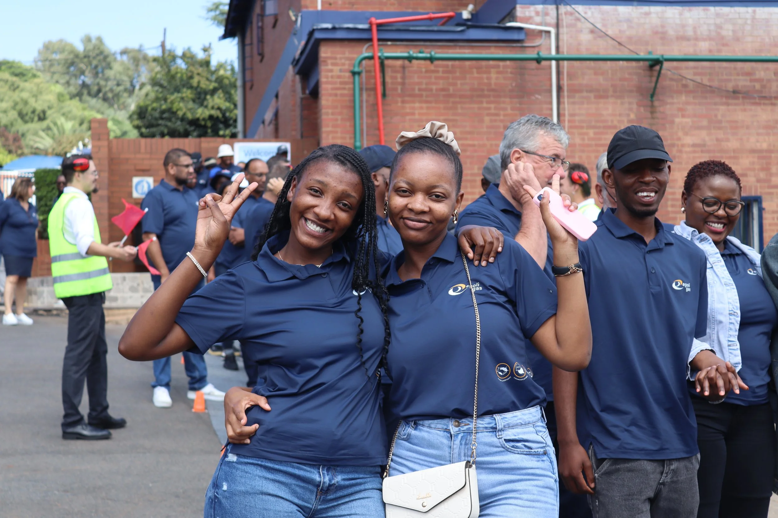 Smiling young women in blue shirts at an outdoor event, with others in the background and a brick building behind them.