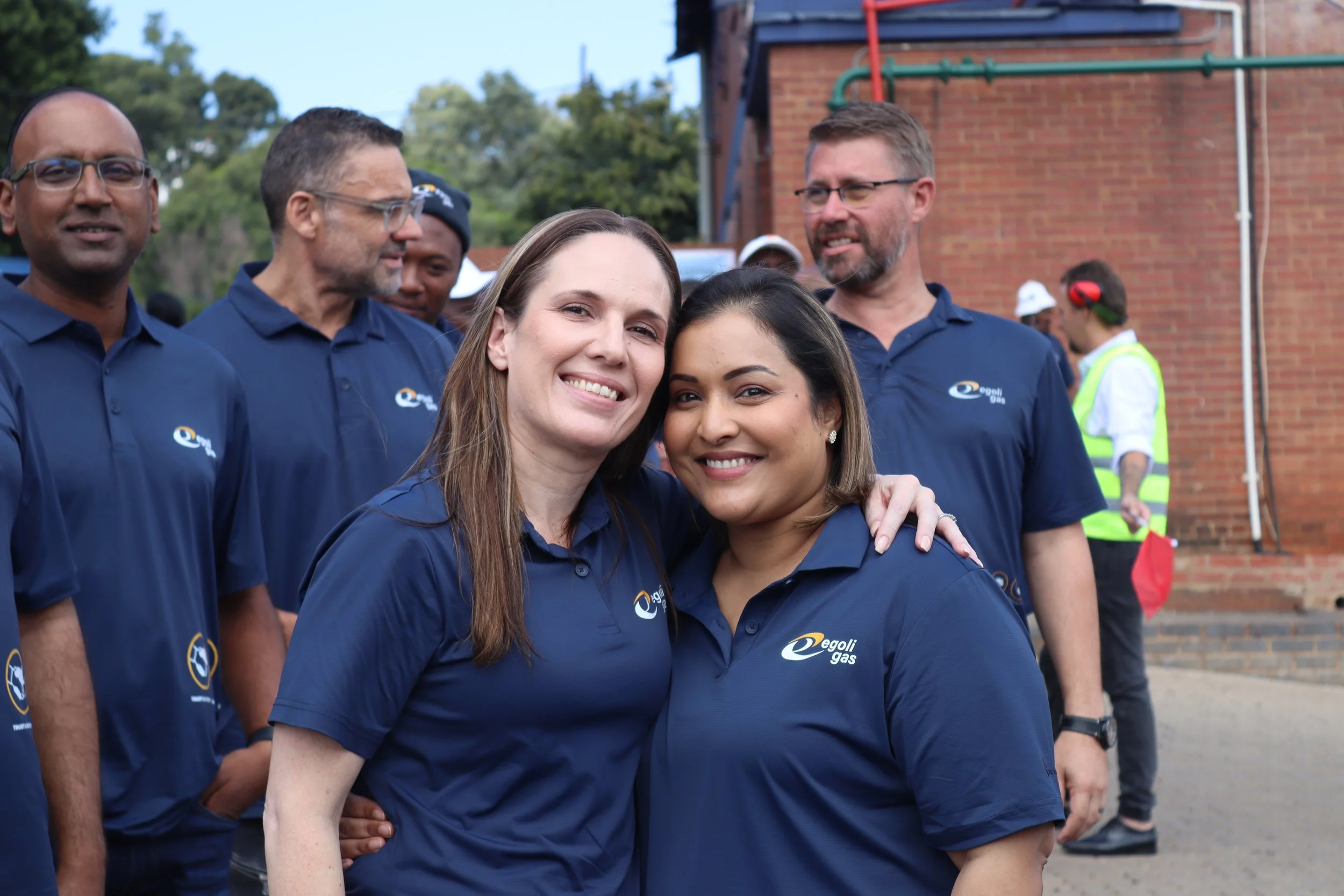 A group of people wearing matching blue shirts with a company logo, gathered outdoors, smiling, with two women in the foreground hugging each other.