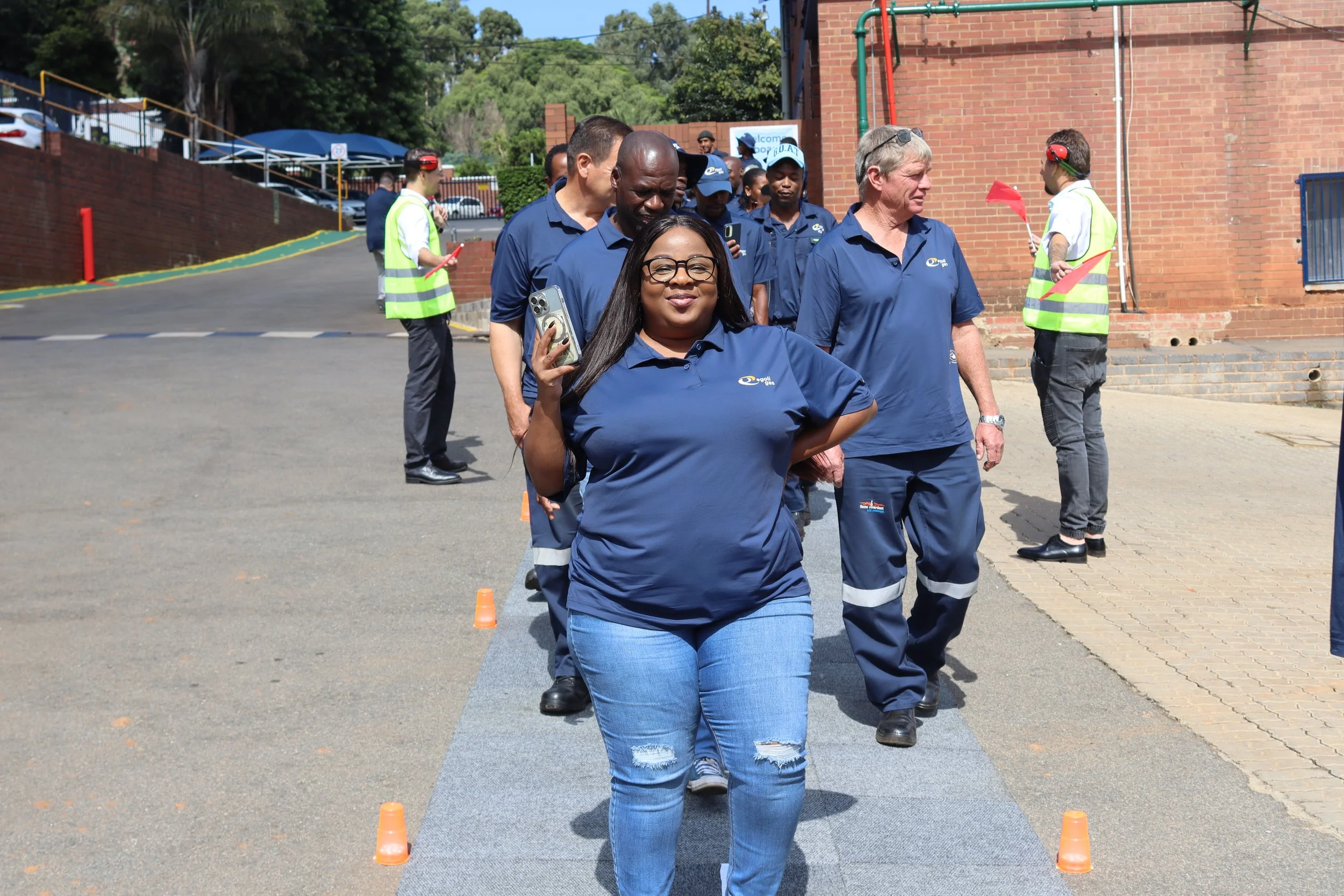 A woman in sunglasses smiling while walking in front of a group of people, some in blue shirts, at an outdoor event near brick buildings and orange cones.