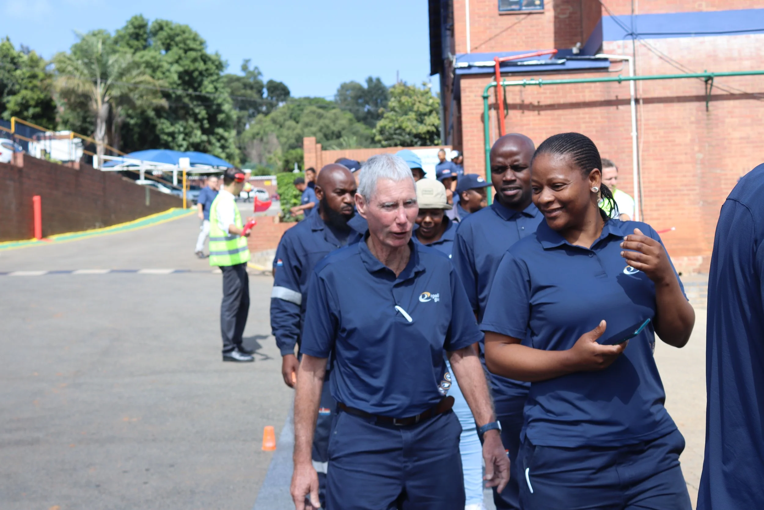 A group of people walking outdoors near a brick building, with trees and parked cars in the background. The group is dressed in dark blue shirts, some of which have a logo. The scene suggests they might be attending a team event or tour.