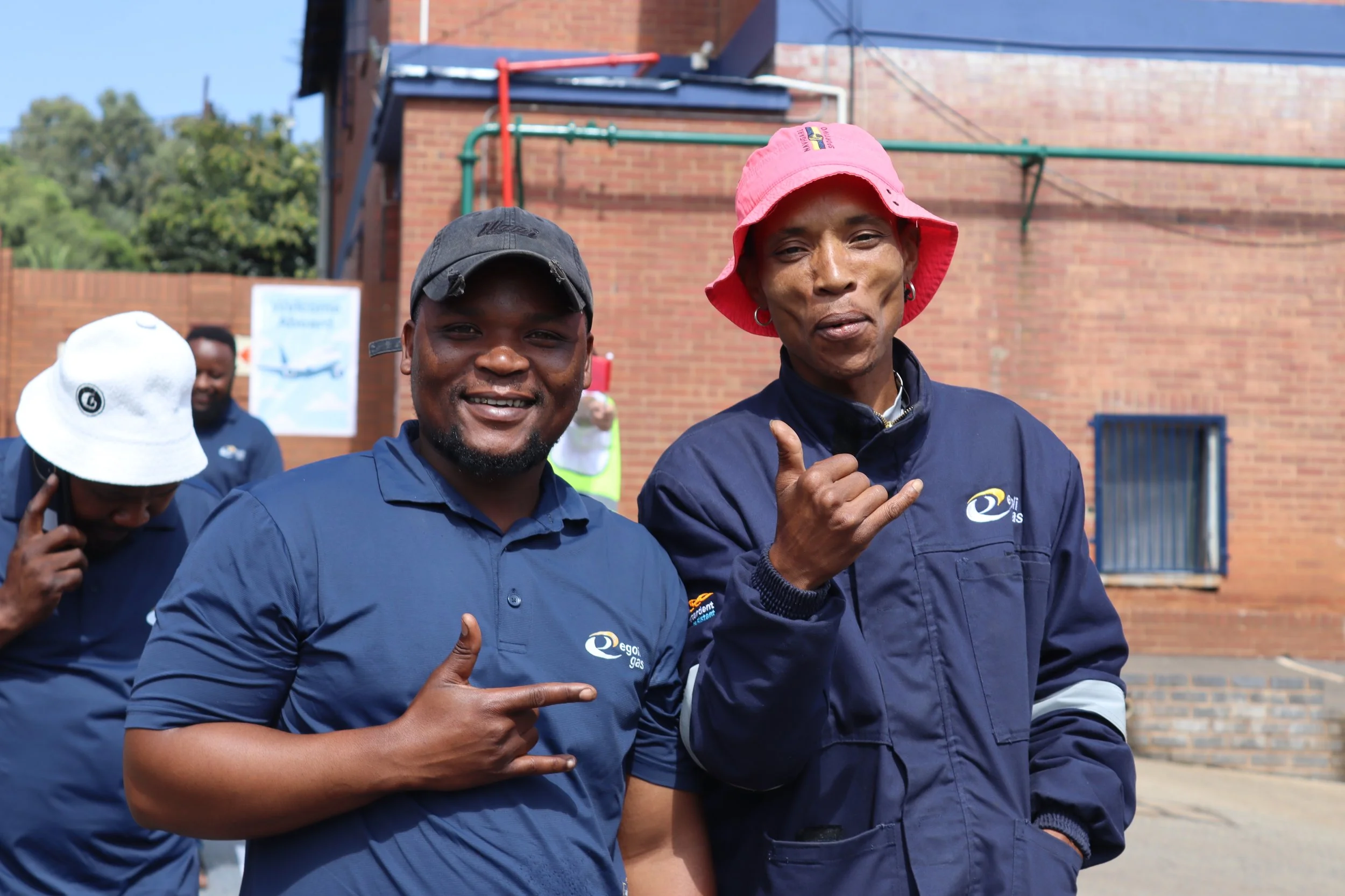 Two men in dark blue jackets with logos, standing outdoors in front of a brick building, smiling and making hand gestures for 'rock on.' One wears a pink bucket hat, the other a black baseball cap. Other people are visible in the background.