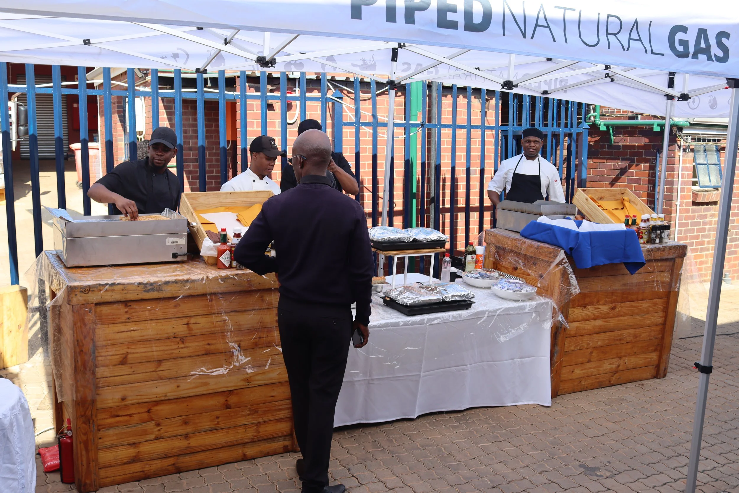 Food stall at an outdoor event selling natural gases, with three workers and a customer.
