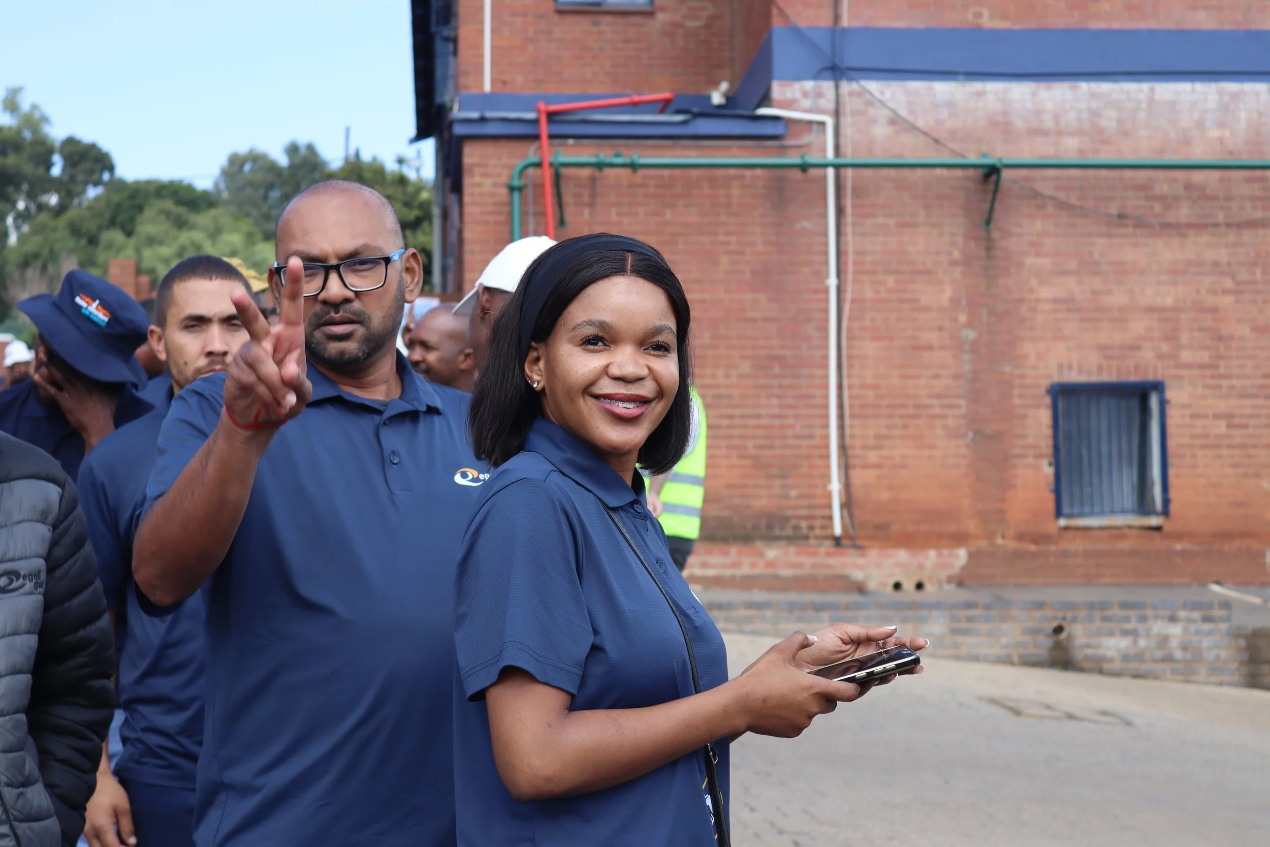 A group of people, wearing dark blue shirts, standing outdoors in front of a brick wall. One woman is smiling and holding a smartphone, while a man next to her is making a peace sign with his fingers.