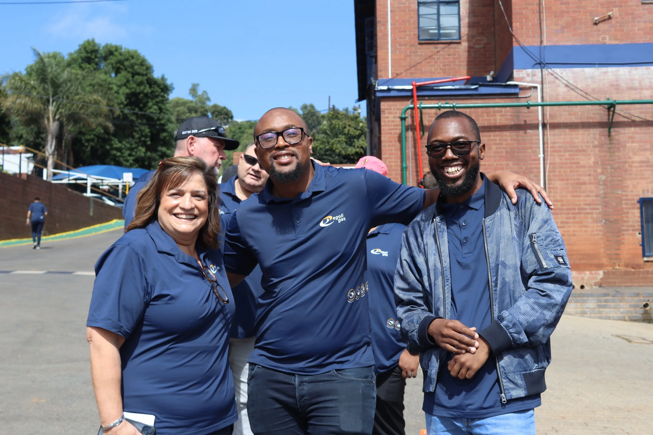 A group of four smiling people wearing blue shirts with a logo, standing outdoors in front of a brick building. Two of them are men with glasses, one woman with brown hair, and a man with a beard wearing a cap and sunglasses. They appear to be at a c
