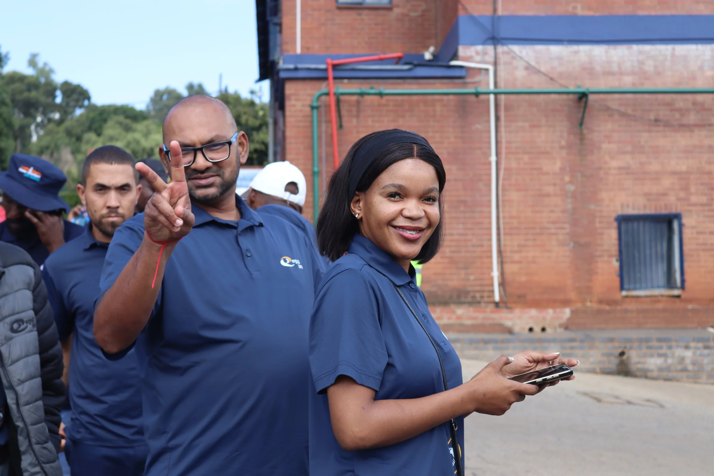 A group of people standing outdoors, with a woman in the foreground holding a smartphone and smiling, and a man behind her making a peace sign with his fingers.
