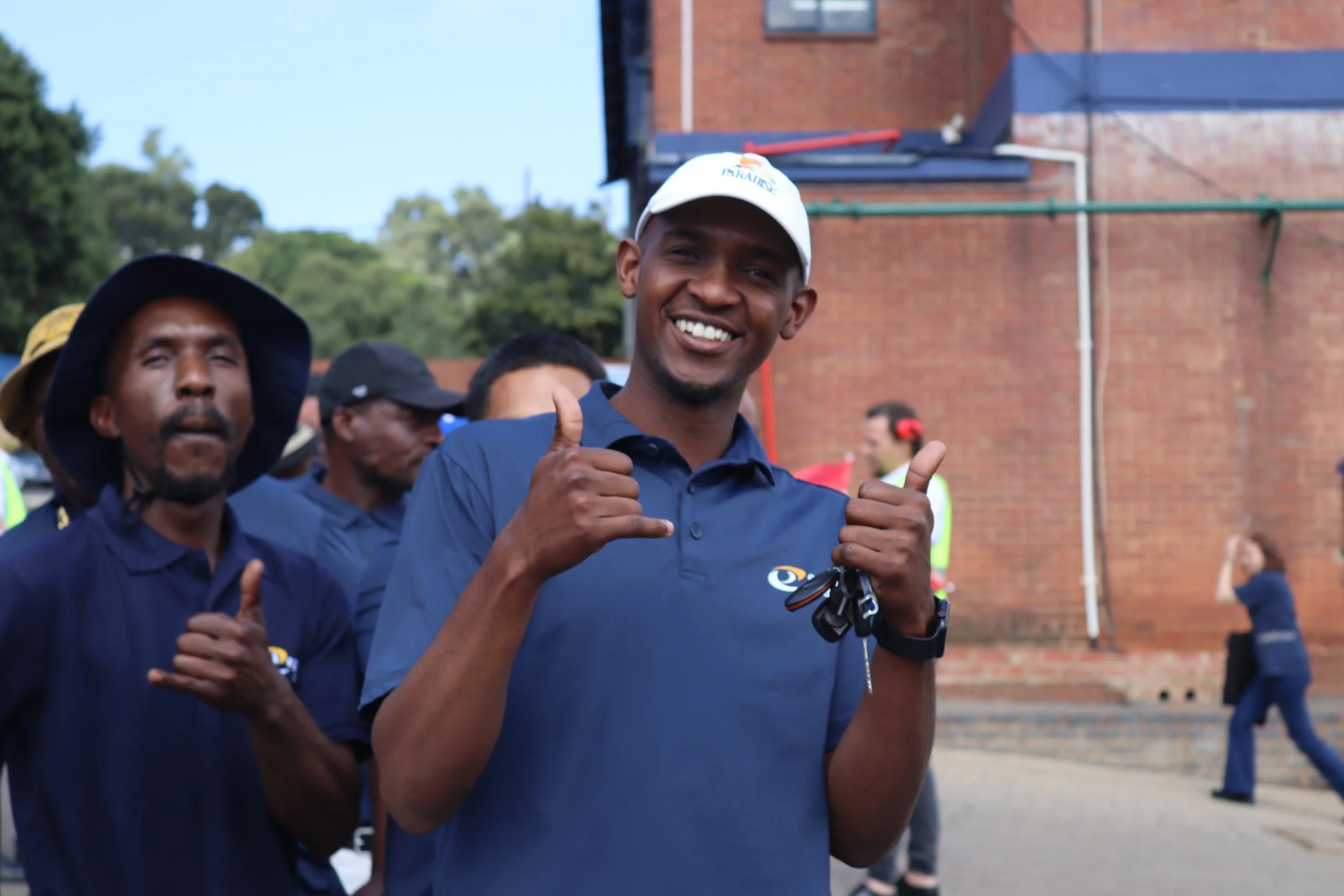 Smiling man wearing a white cap giving a thumbs-up at an outdoor event, with other people in the background.