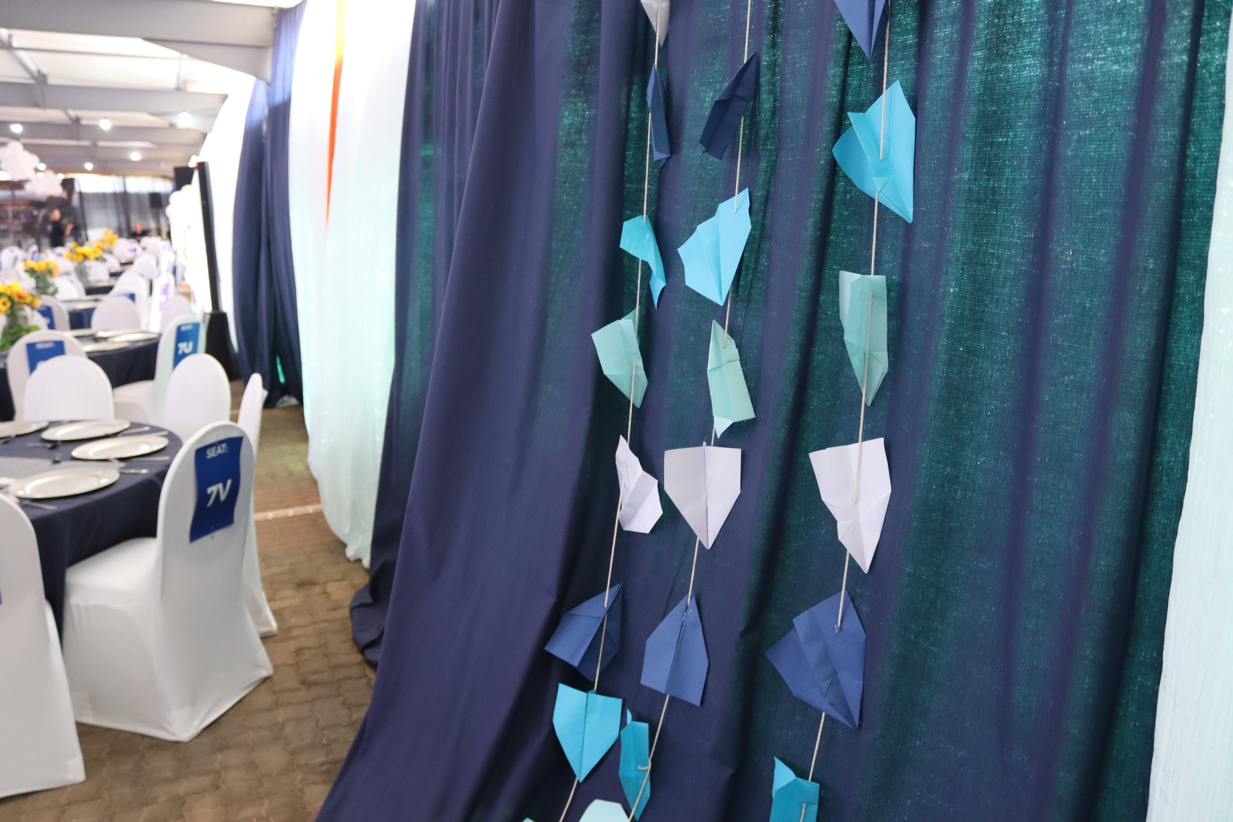 Decorative display of blue and white paper umbrellas hanging on strings on a dark blue curtain, set up at an event with tables and chairs in the background.