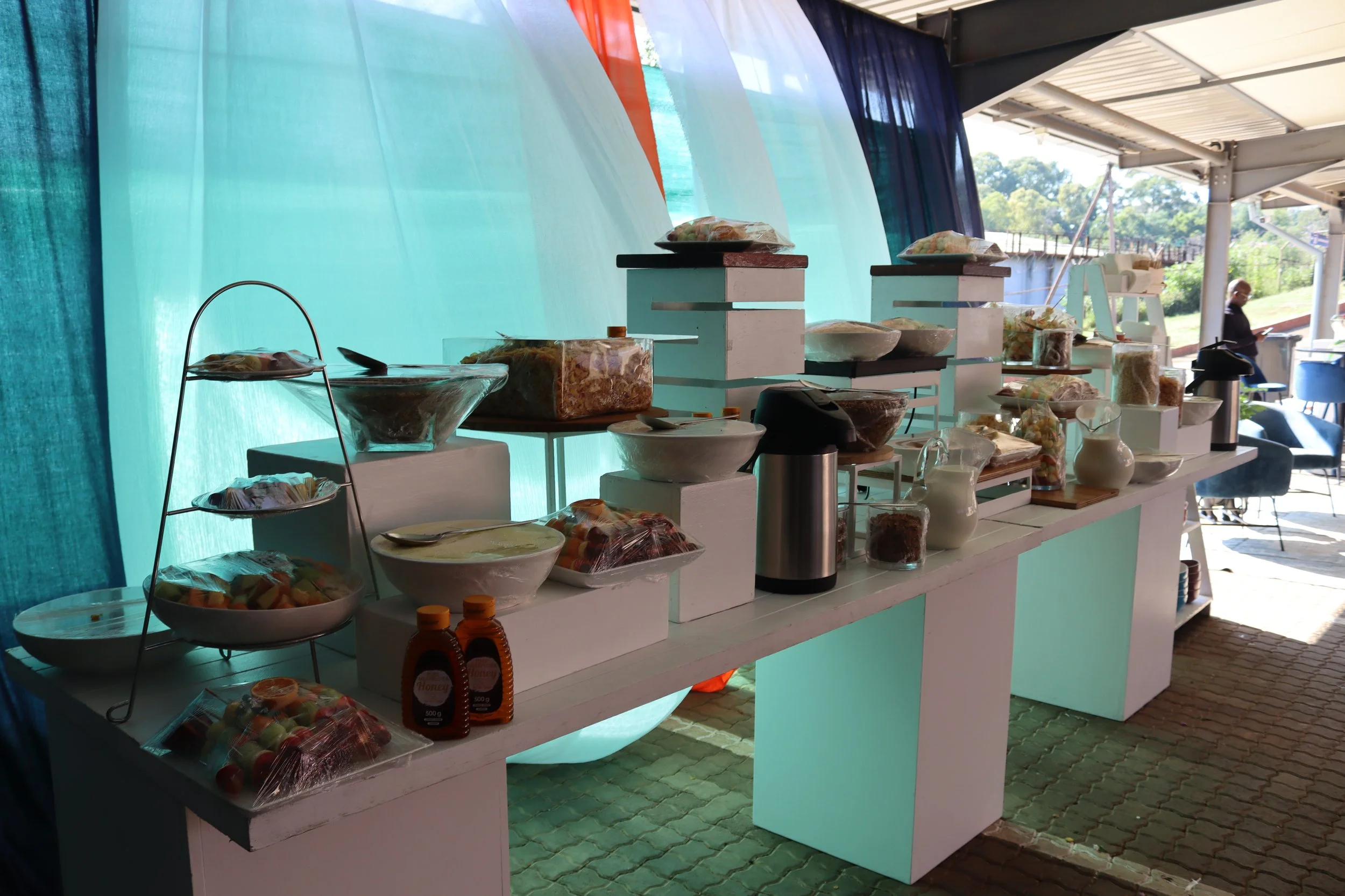 Display of food products on a table under a canopy, with jars, packaged snacks, and bowls of fruit salad, set outdoors on a sunny day.
