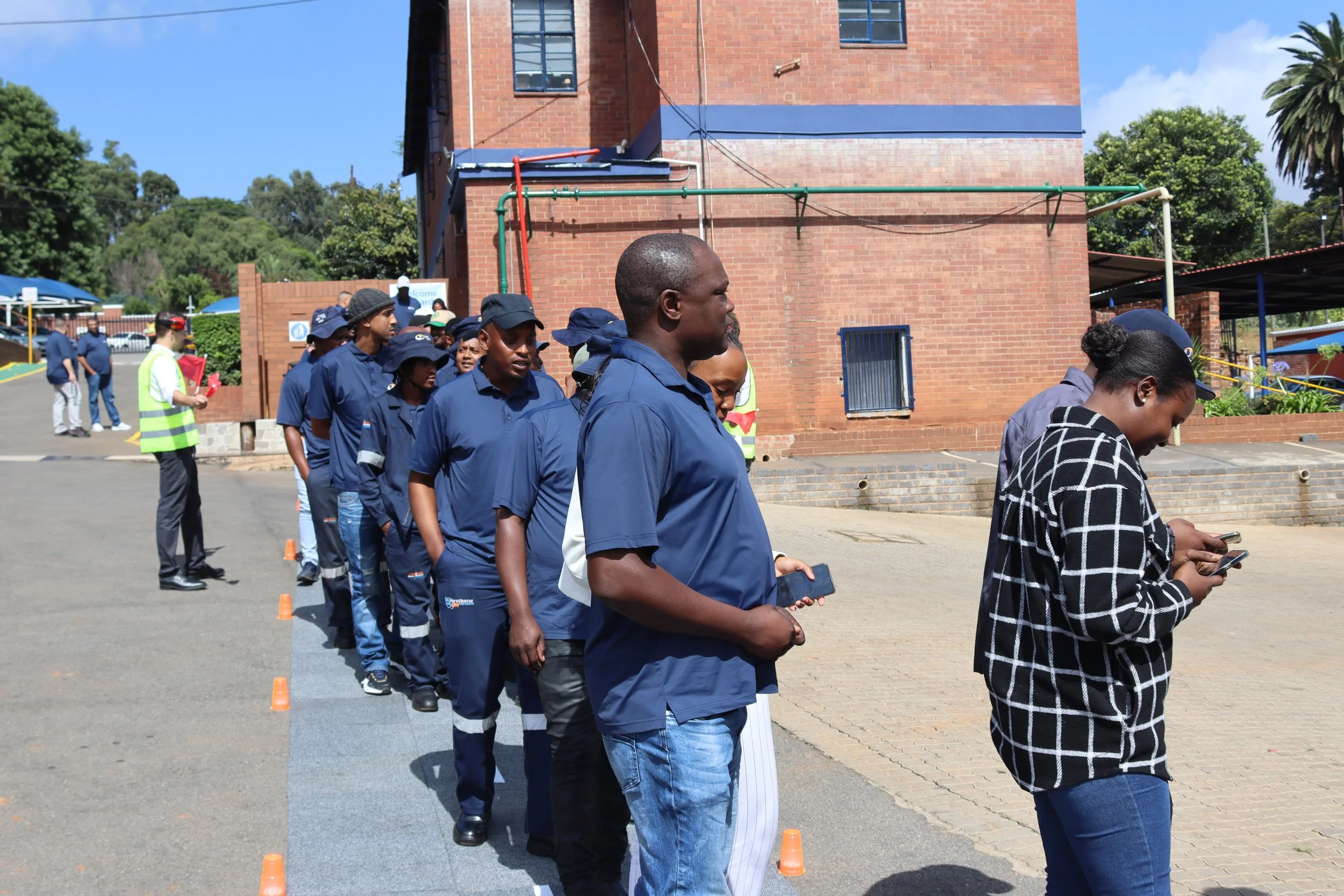 Line of people standing outdoors, some looking at their phones, in front of a brick building with trees in the background, during daytime.