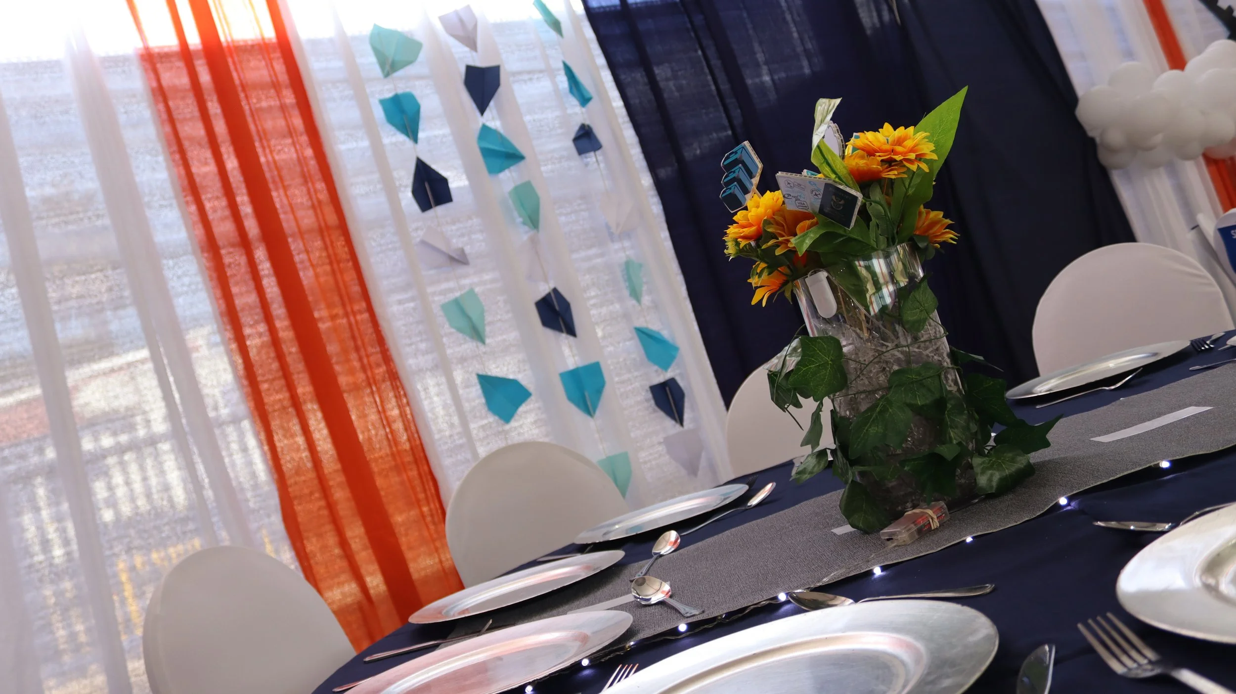 Decorated event table with a centerpiece of yellow flowers and green leaves, surrounded by white chairs, set with plates and utensils, with a backdrop of fabric and paper decorations in shades of blue, white, and orange.