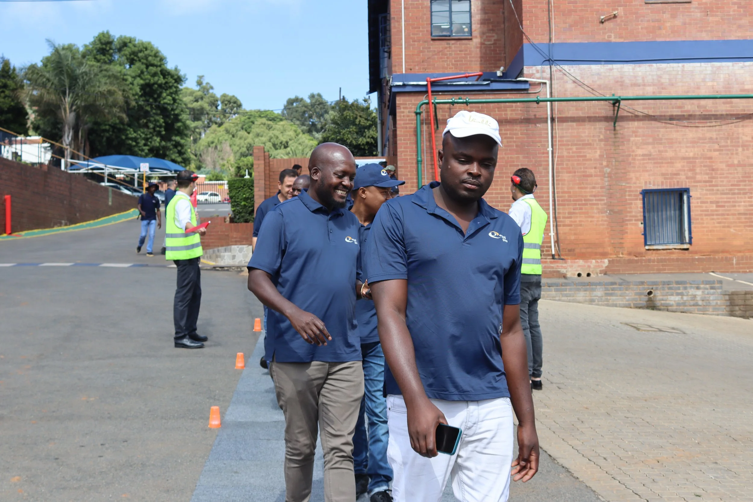 Group of people walking outdoors along a line of orange cones, with a brick building and trees in the background. Some individuals are wearing blue shirts with a logo, and others are wearing safety vests.