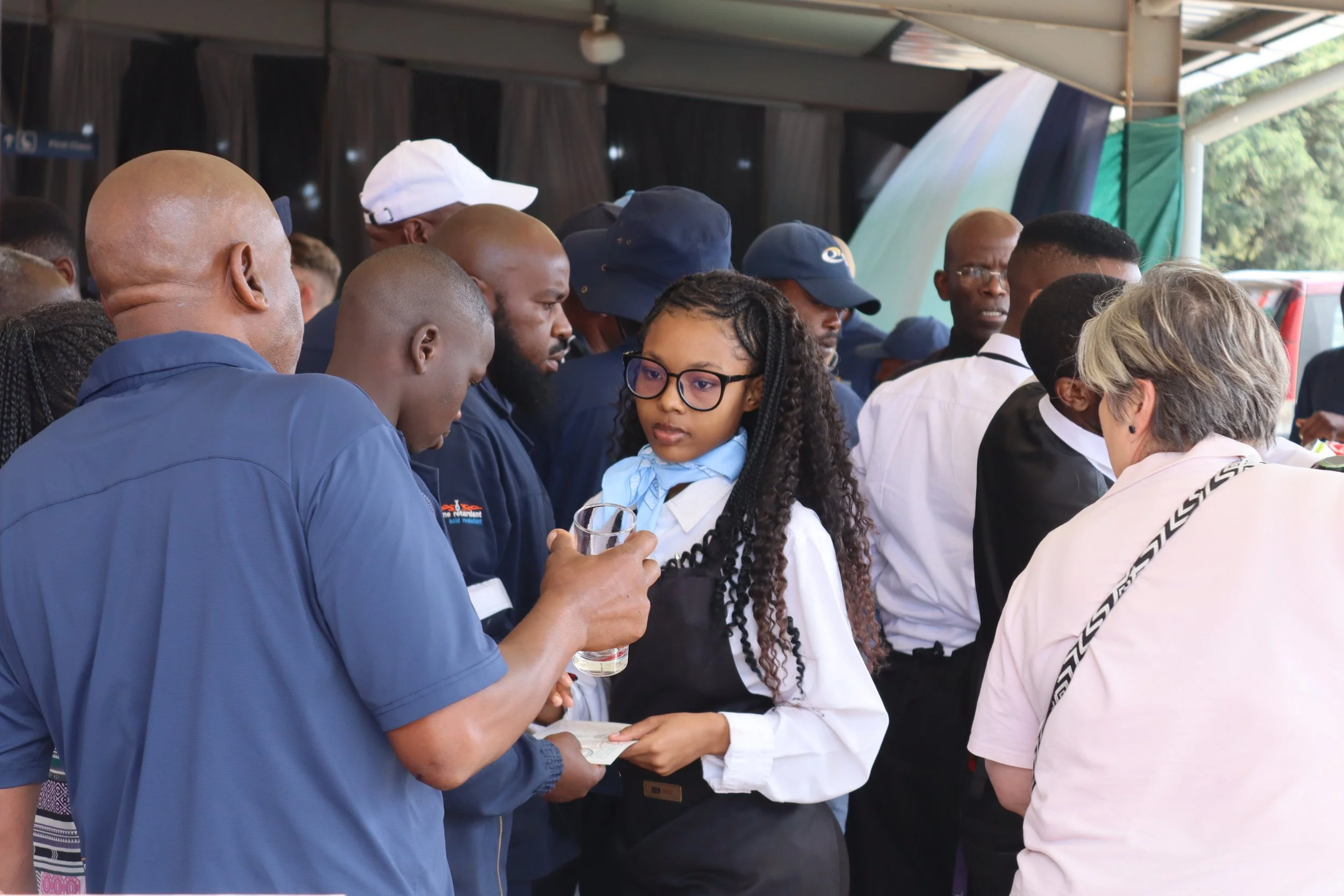 Group of people gathered, with some looking at papers and one person holding a glass. The scene appears to involve a discussion or sharing of information.