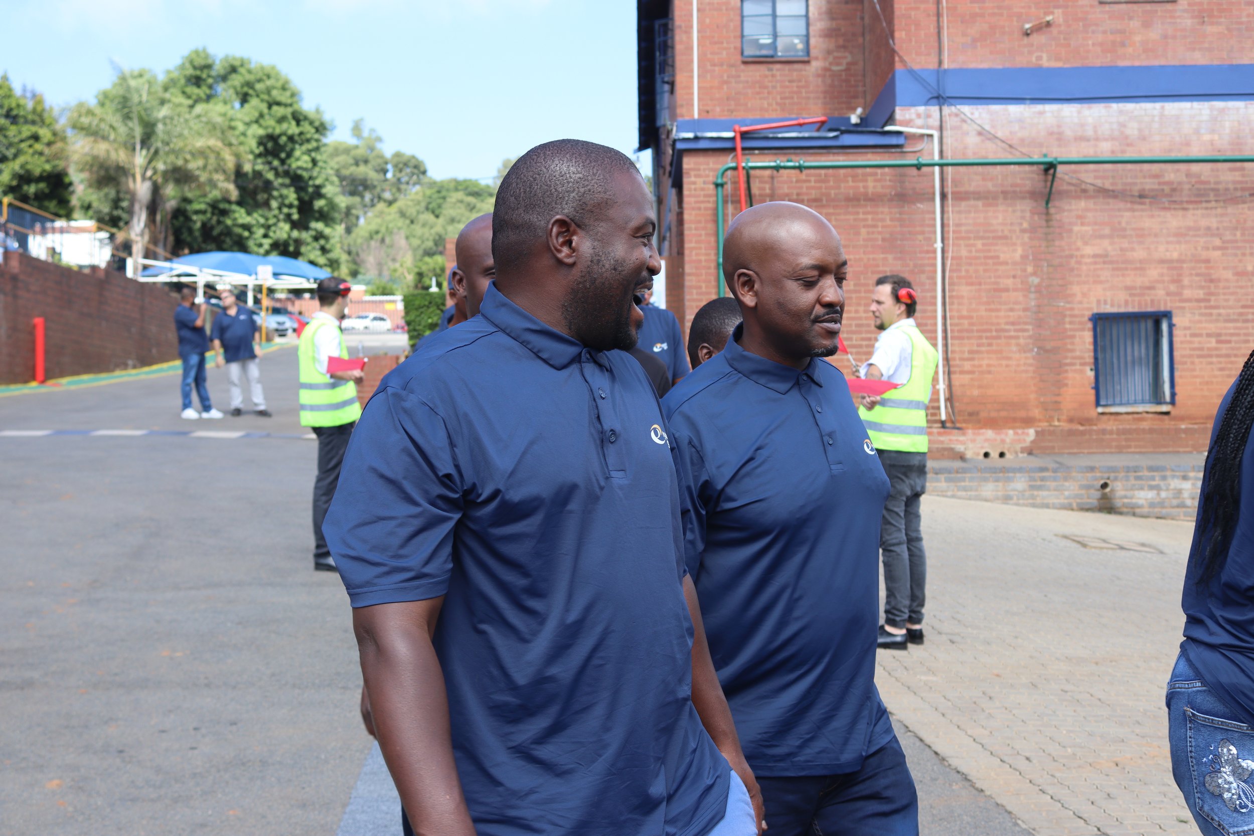 Two men in blue shirts standing outdoors near a brick building, smiling and engaging with each other, with other people in the background.
