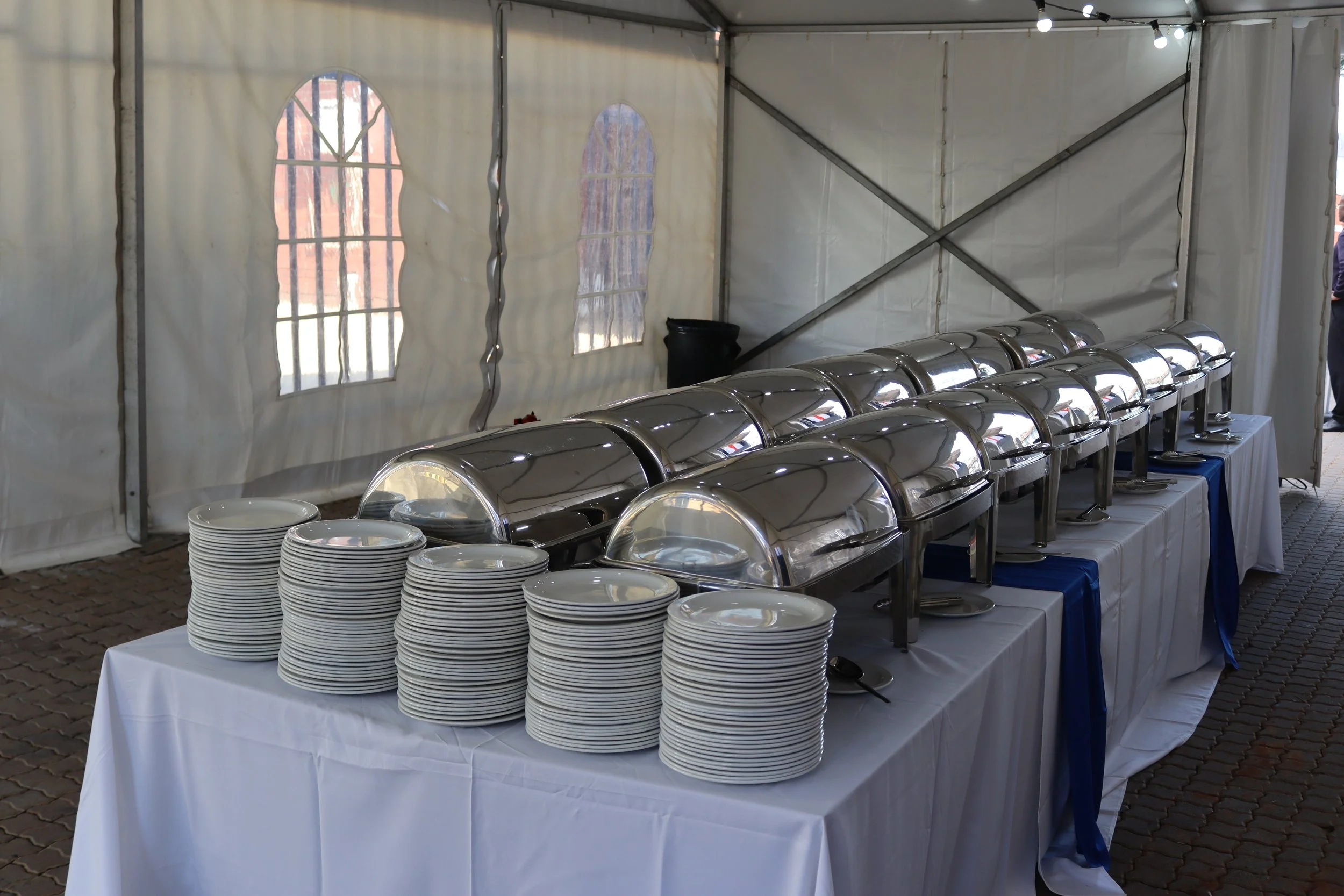A buffet setup inside a white tent with stacks of plates on a table and chafing dishes for serving food.