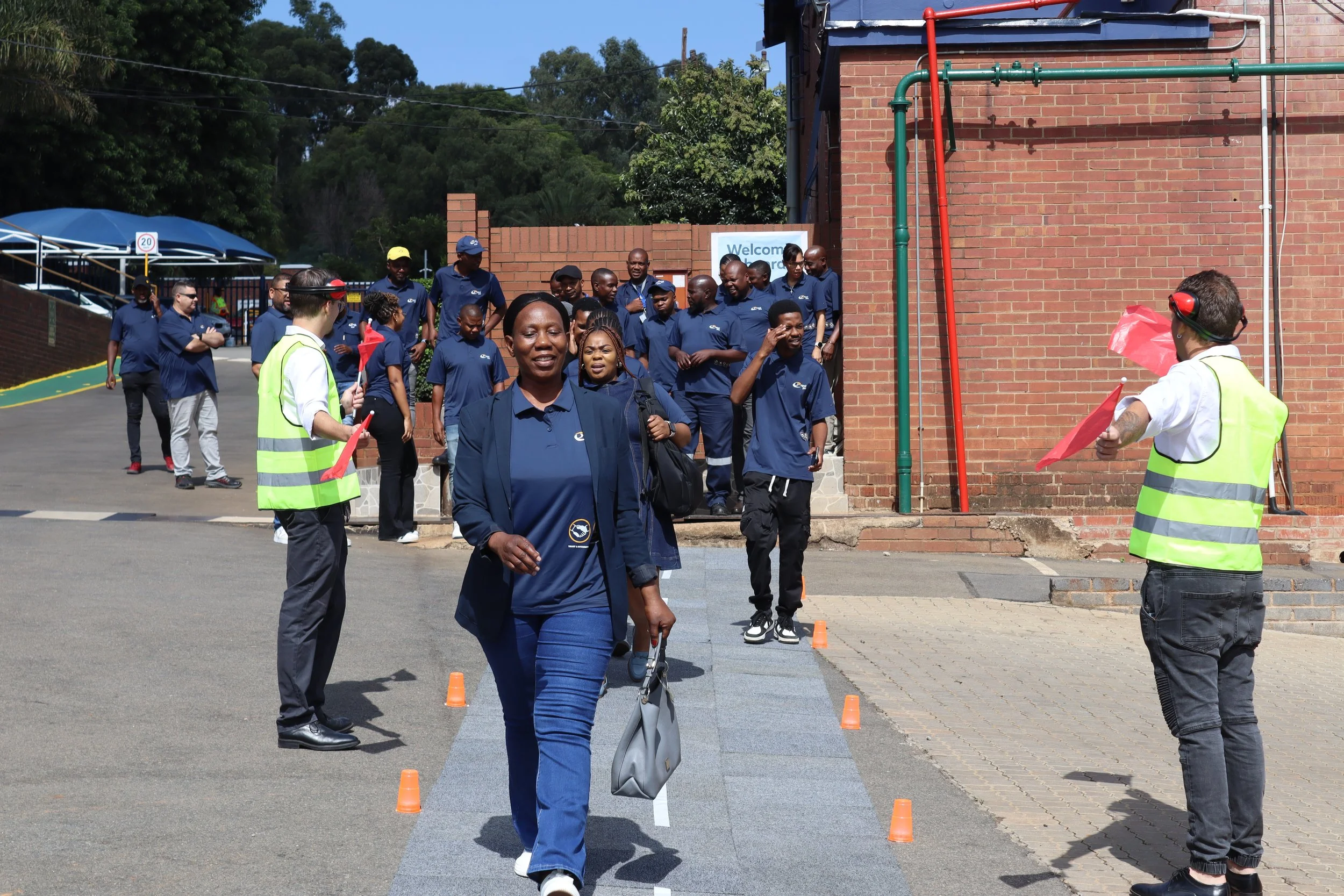 Group of people, mostly in navy uniforms, walking outside near brick buildings and trees on a sunny day.